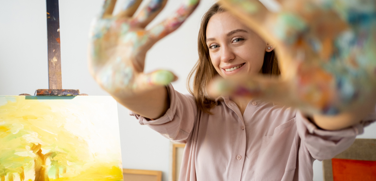 Woman with paint-covered hands smiles at the camera, framing the canvas on an easel in an art studio.