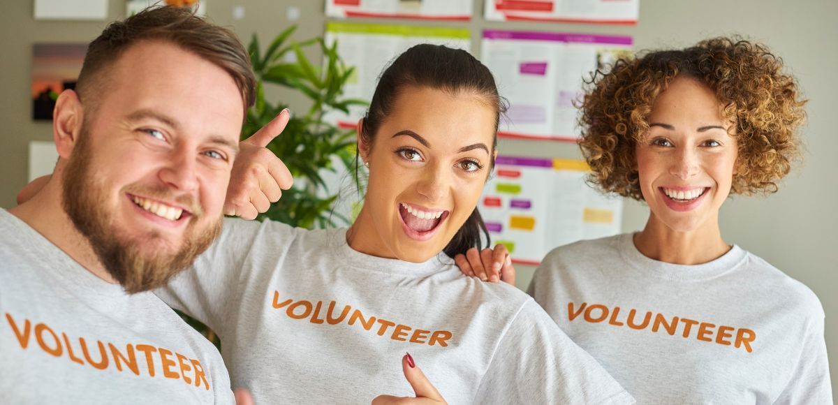 Three smiling volunteers in gray “VOLUNTEER” shirts standing together indoors.
