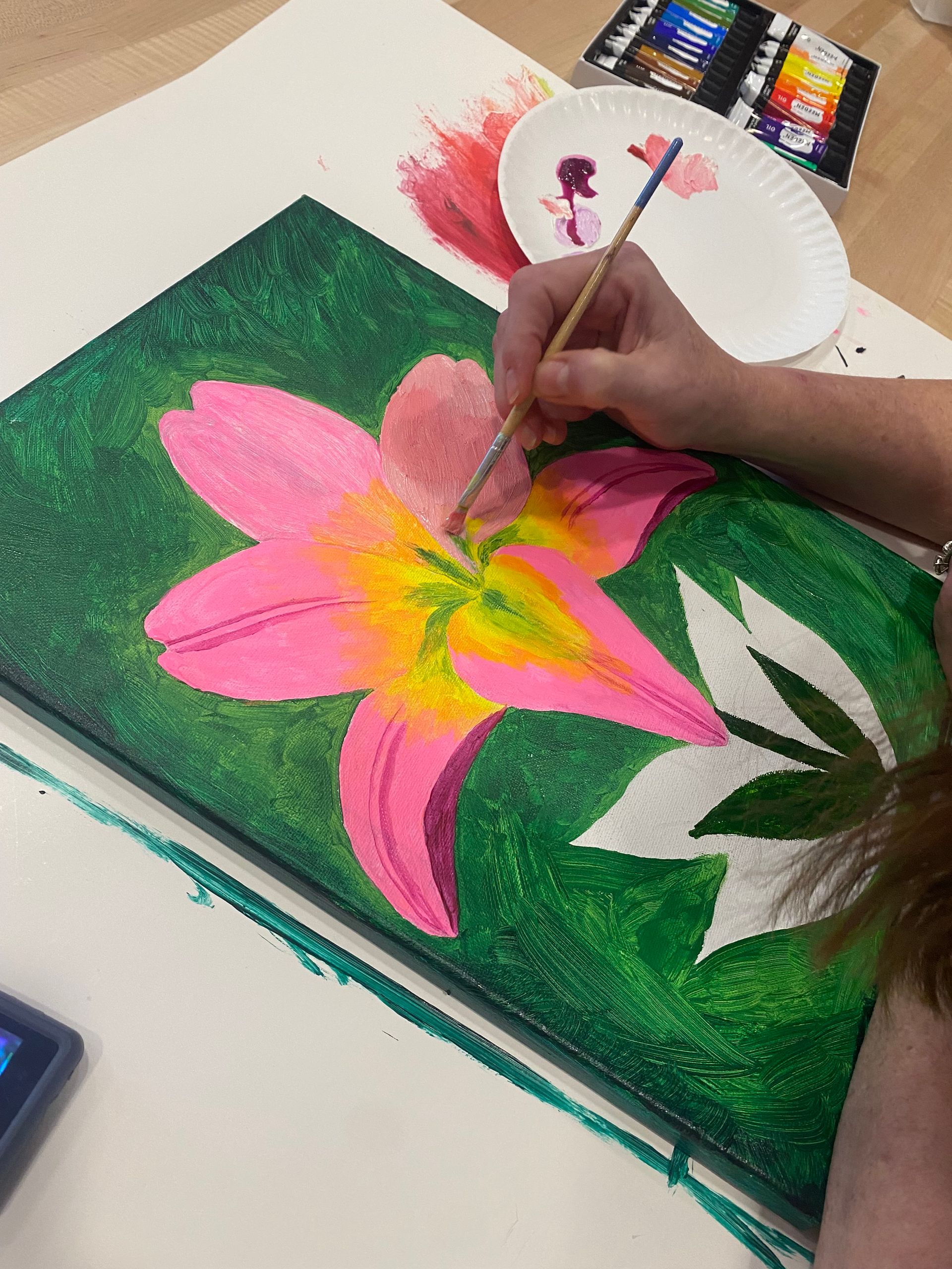 Person painting a pink lily on a green background. Brush in hand, applying paint with a palette nearby.