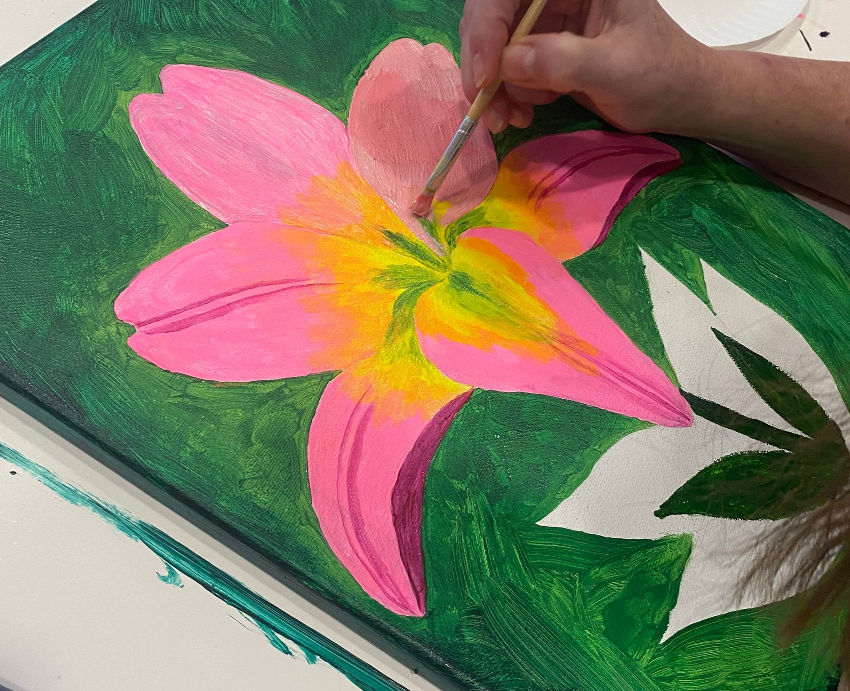 Person painting a pink and yellow lily on a green canvas with a brush.