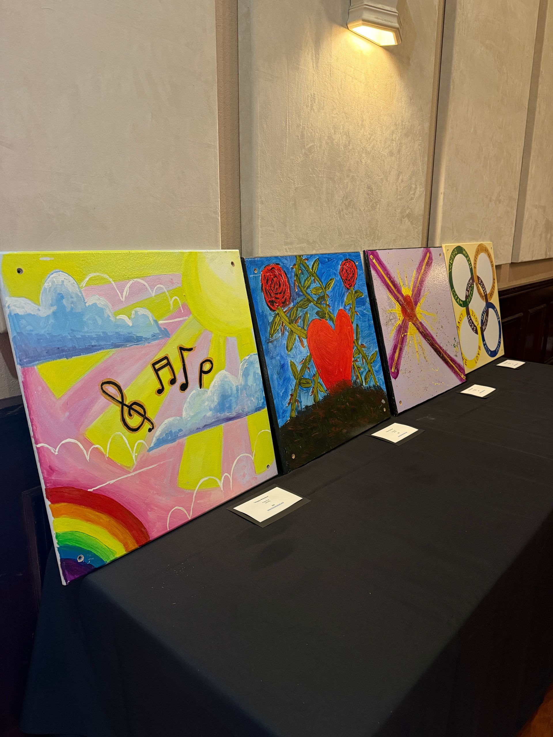 Four colorful square paintings displayed on a black table, featuring musical, floral, heart, and Olympic ring motifs.