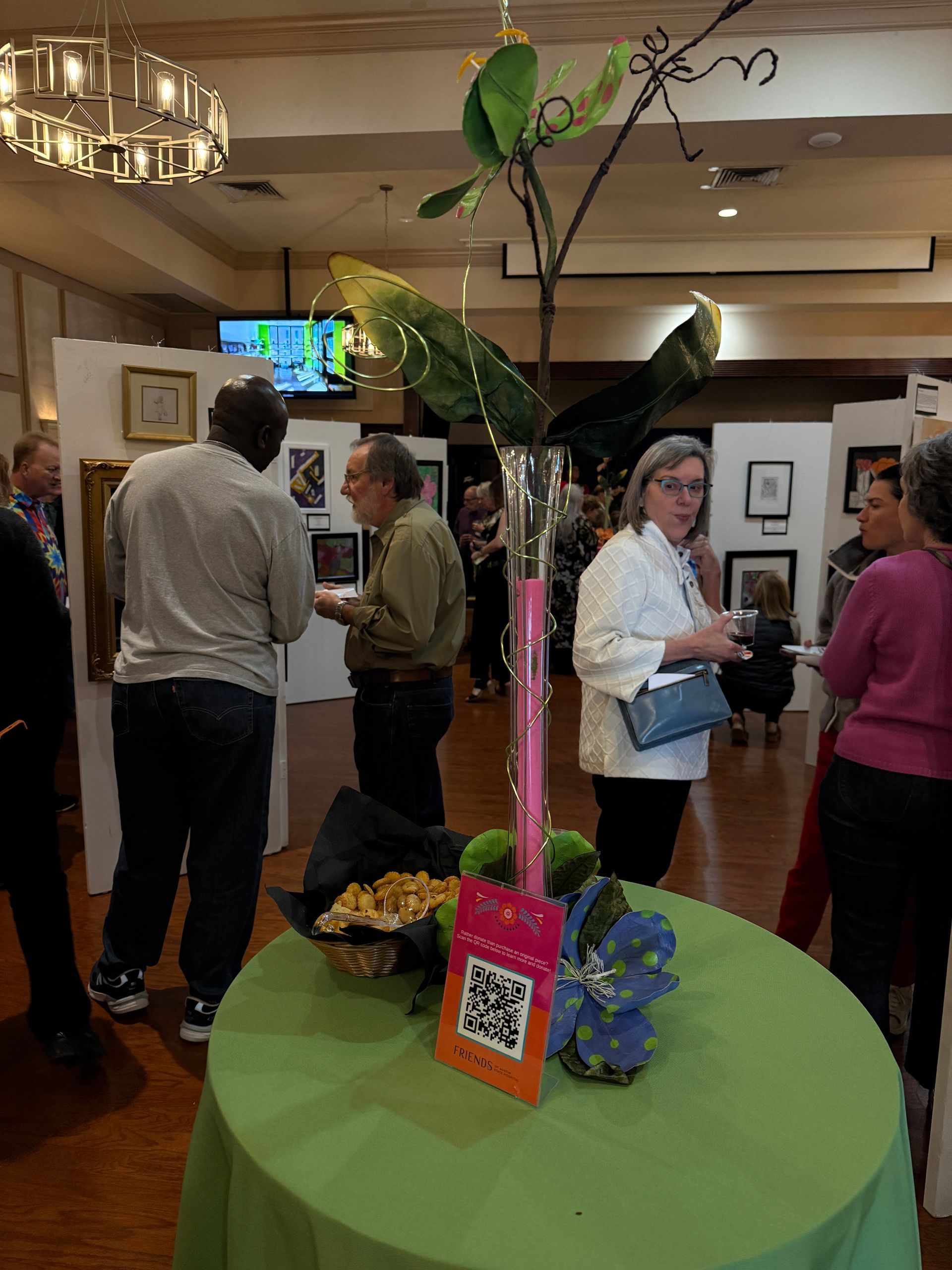 Attendees mingle at an indoor art gallery event, standing around a green table with a tall decorative centerpiece.