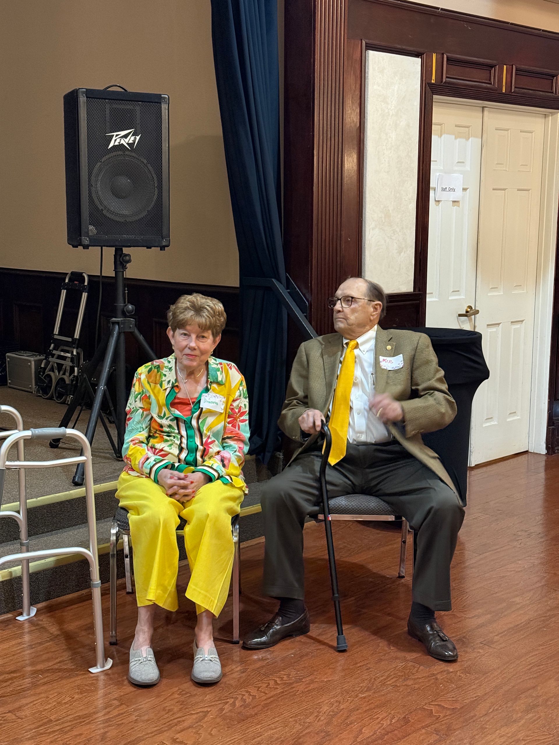 A person in a patterned blazer and yellow pants sits next to a person in a brown blazer and yellow tie with a cane.