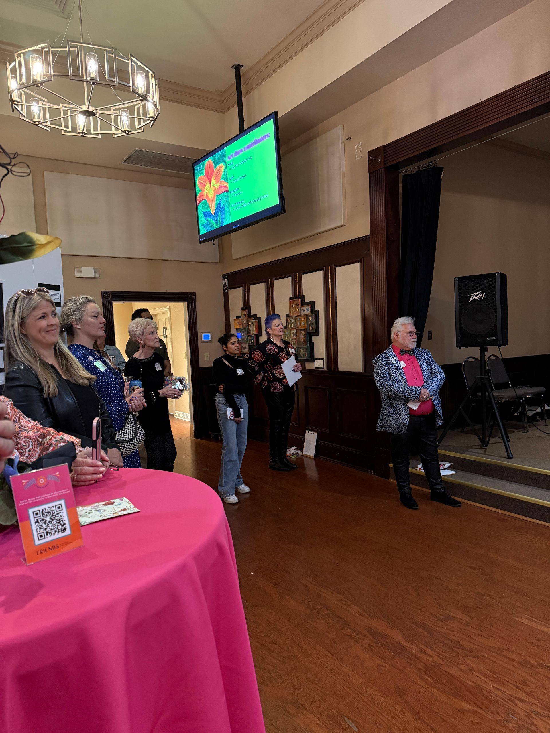 A group of people standing in a hall with a pink-covered table in the foreground and a TV screen displayed on the wall.