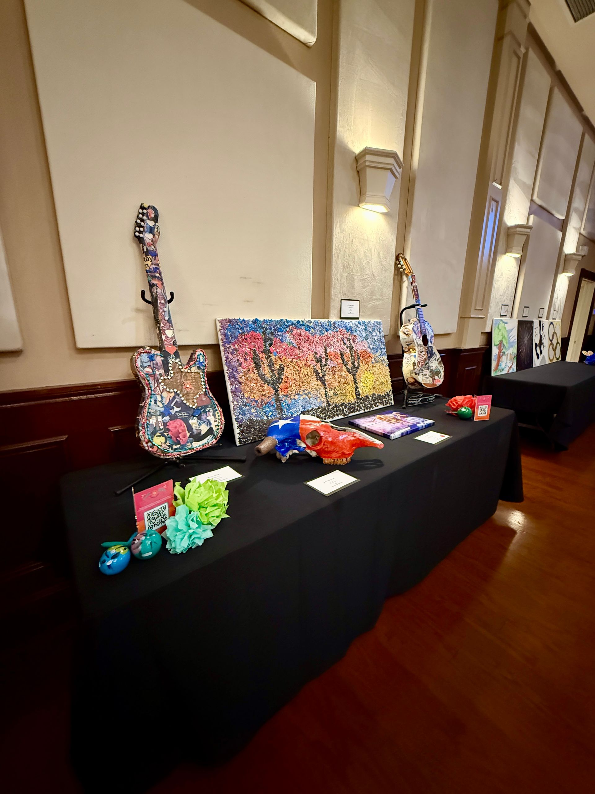 Display table with colorful guitars and art for sale in a well-lit room.