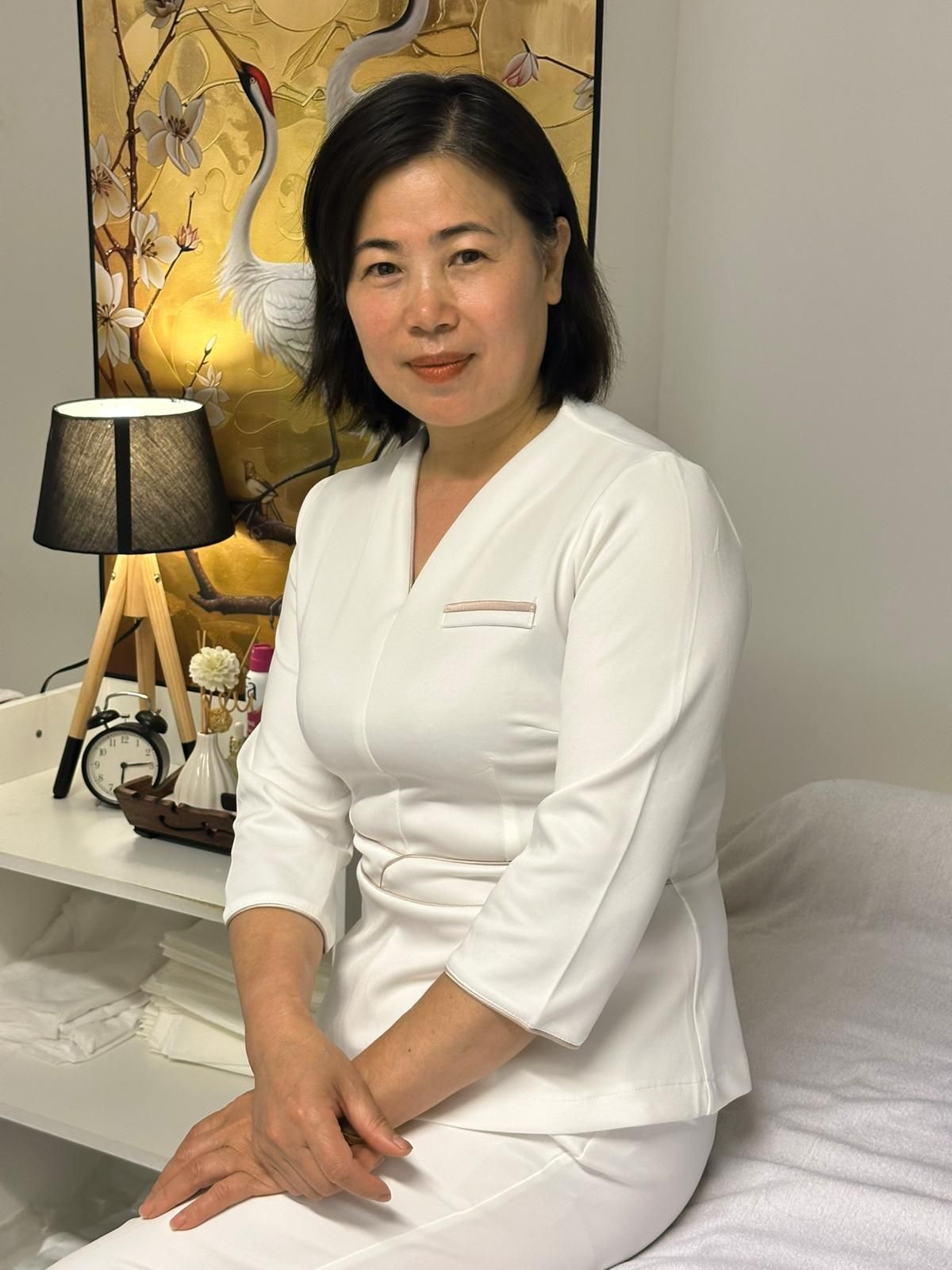 Vrouw in een wit uniform, zittend, in een spa-achtige setting met een gouden kunstwerk aan de muur.