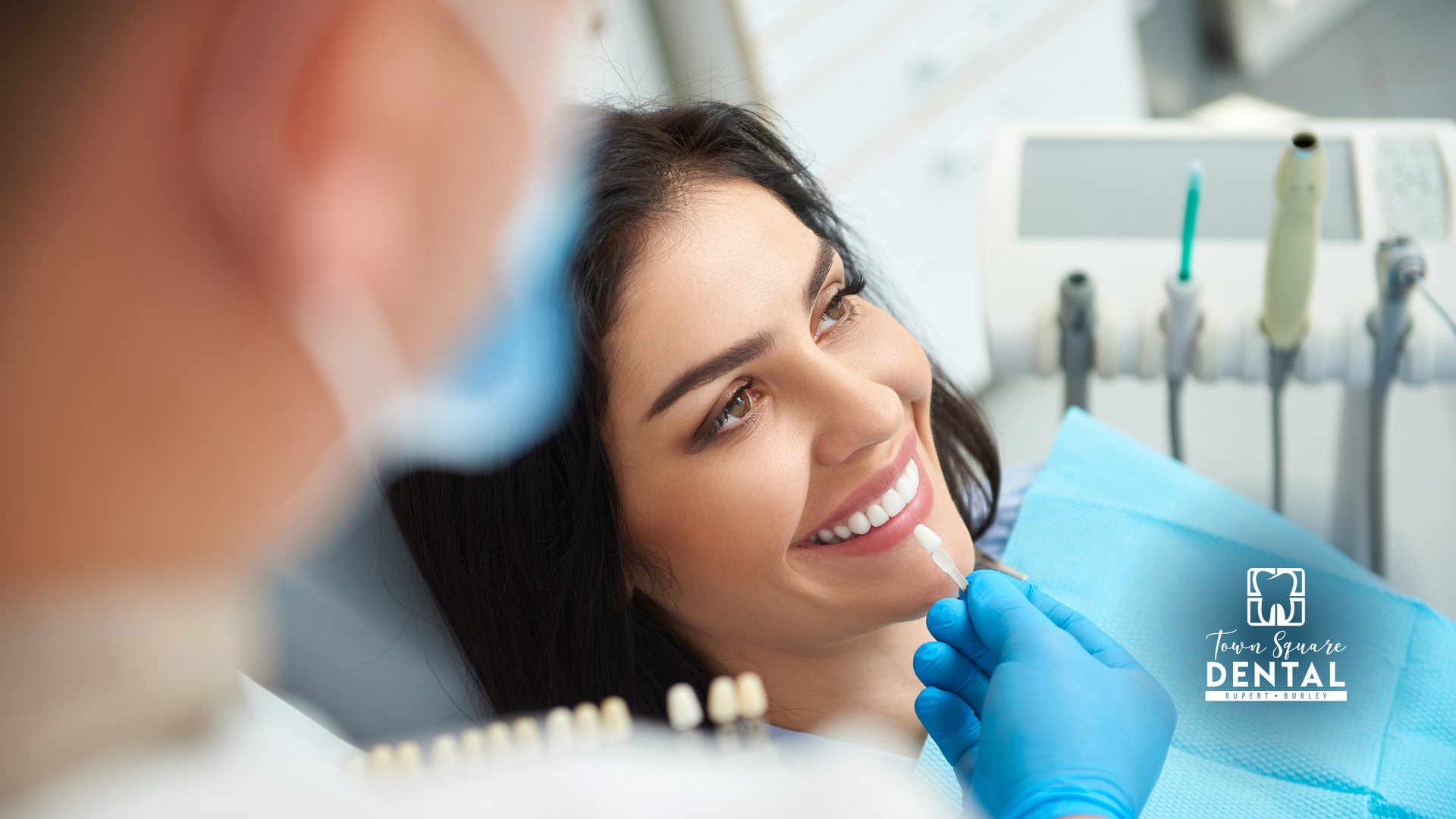 A woman is sitting in a dental chair smiling while a dentist examines her teeth.