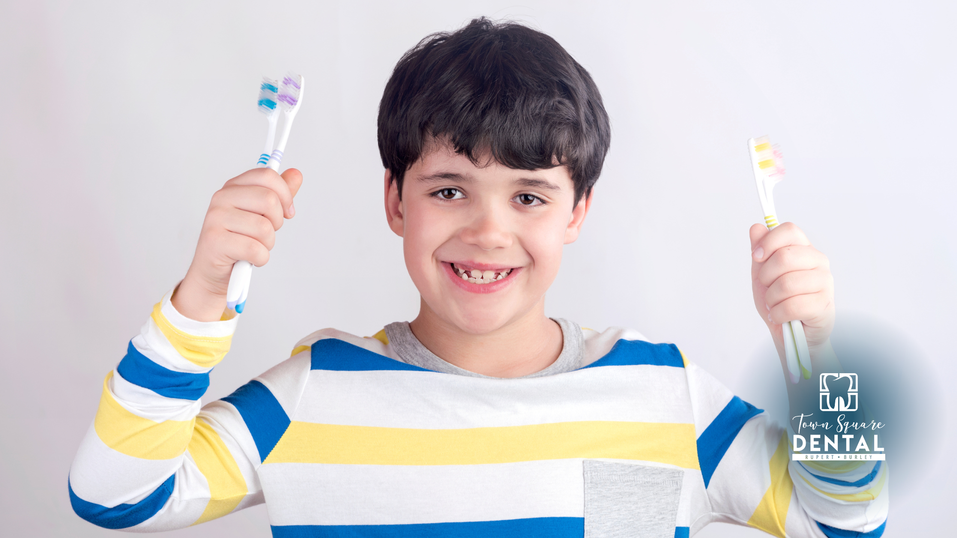 Boy with missing tooth smiles, holding two toothbrushes.