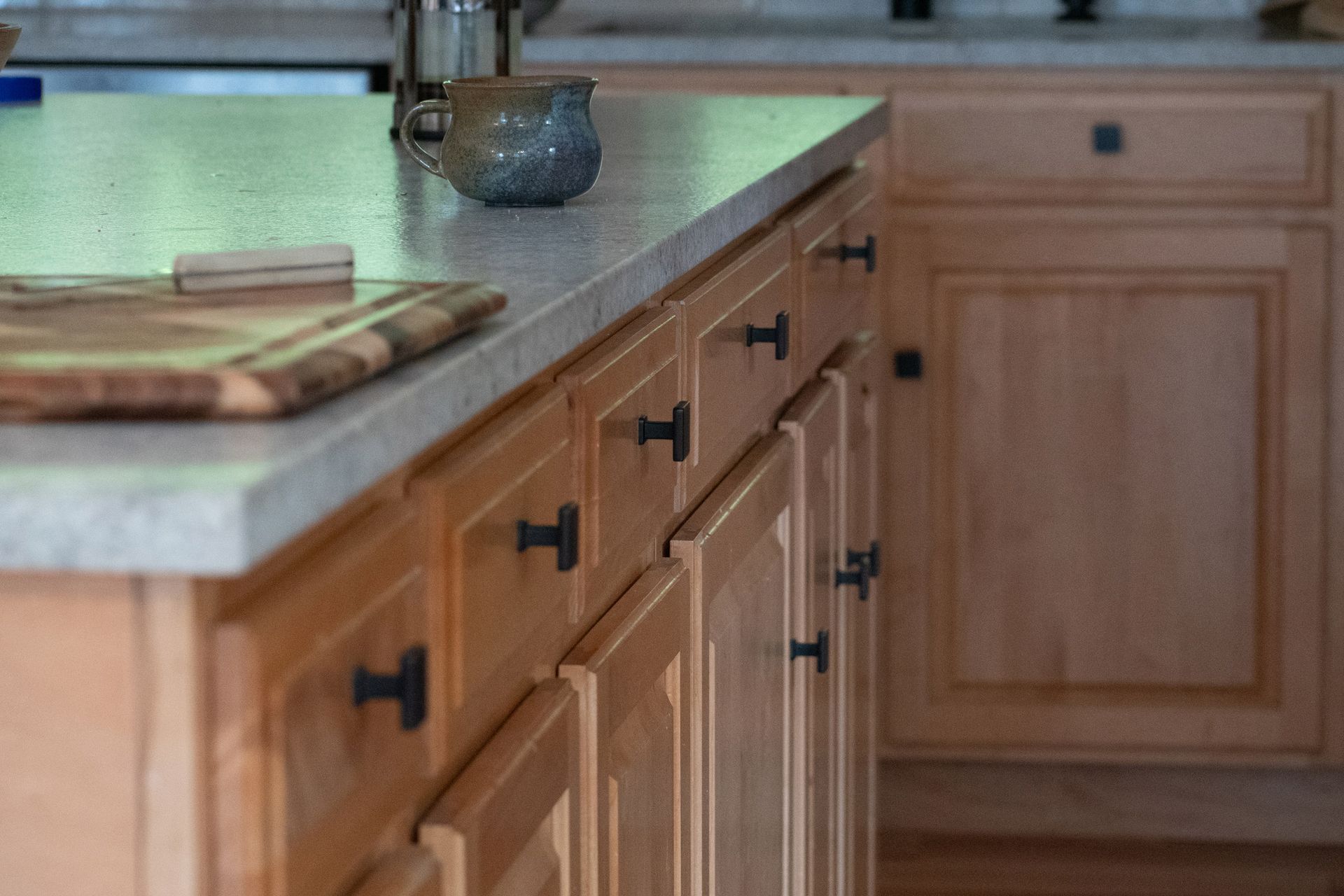 Close-up of kitchen island with light wood cabinets, granite countertop, and black hardware.