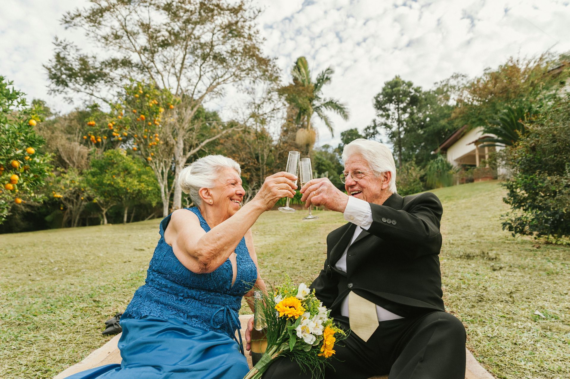 An elderly couple is sitting on a blanket in the grass toasting with champagne.