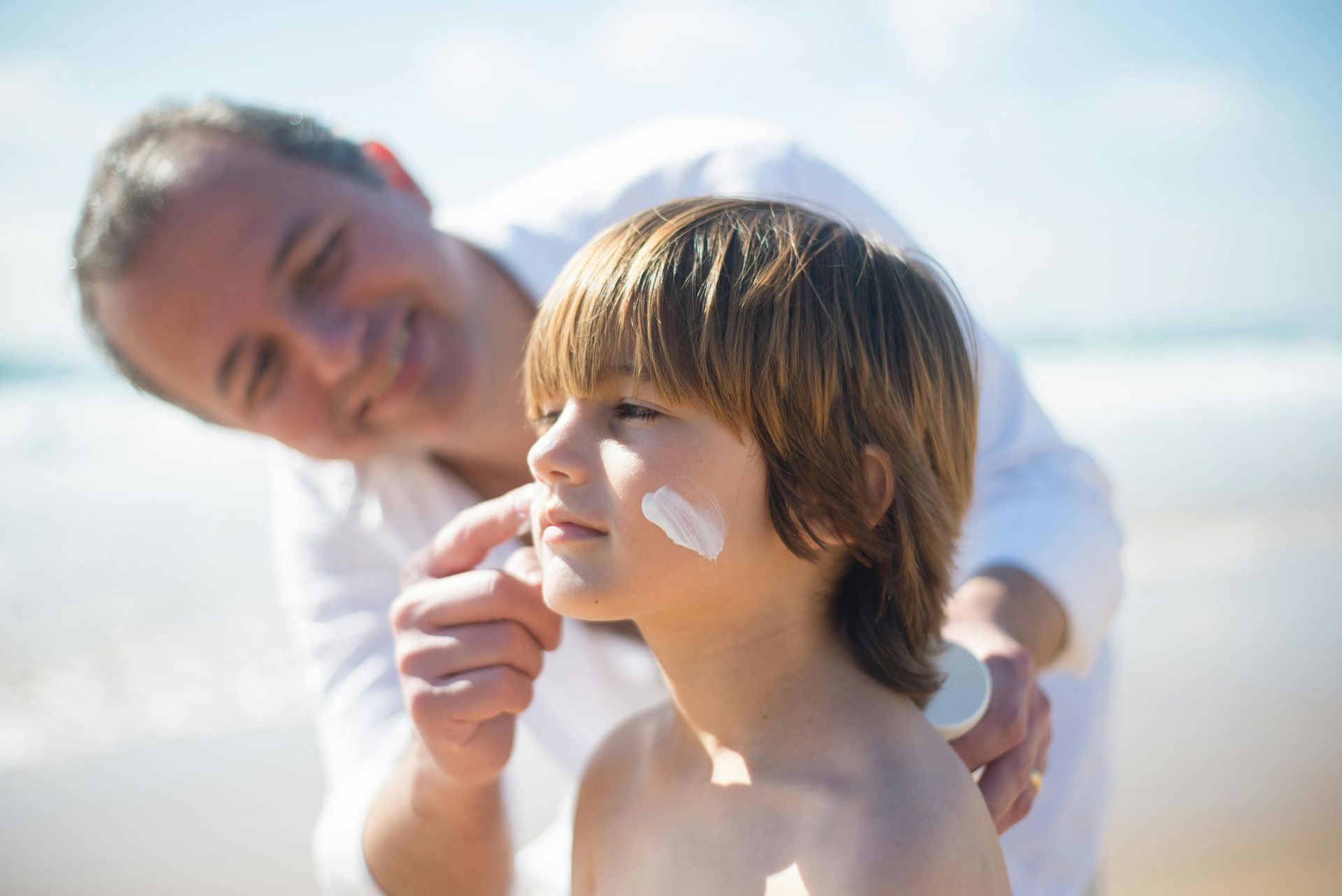 A man is applying sunscreen to a young boy 's face on the beach.