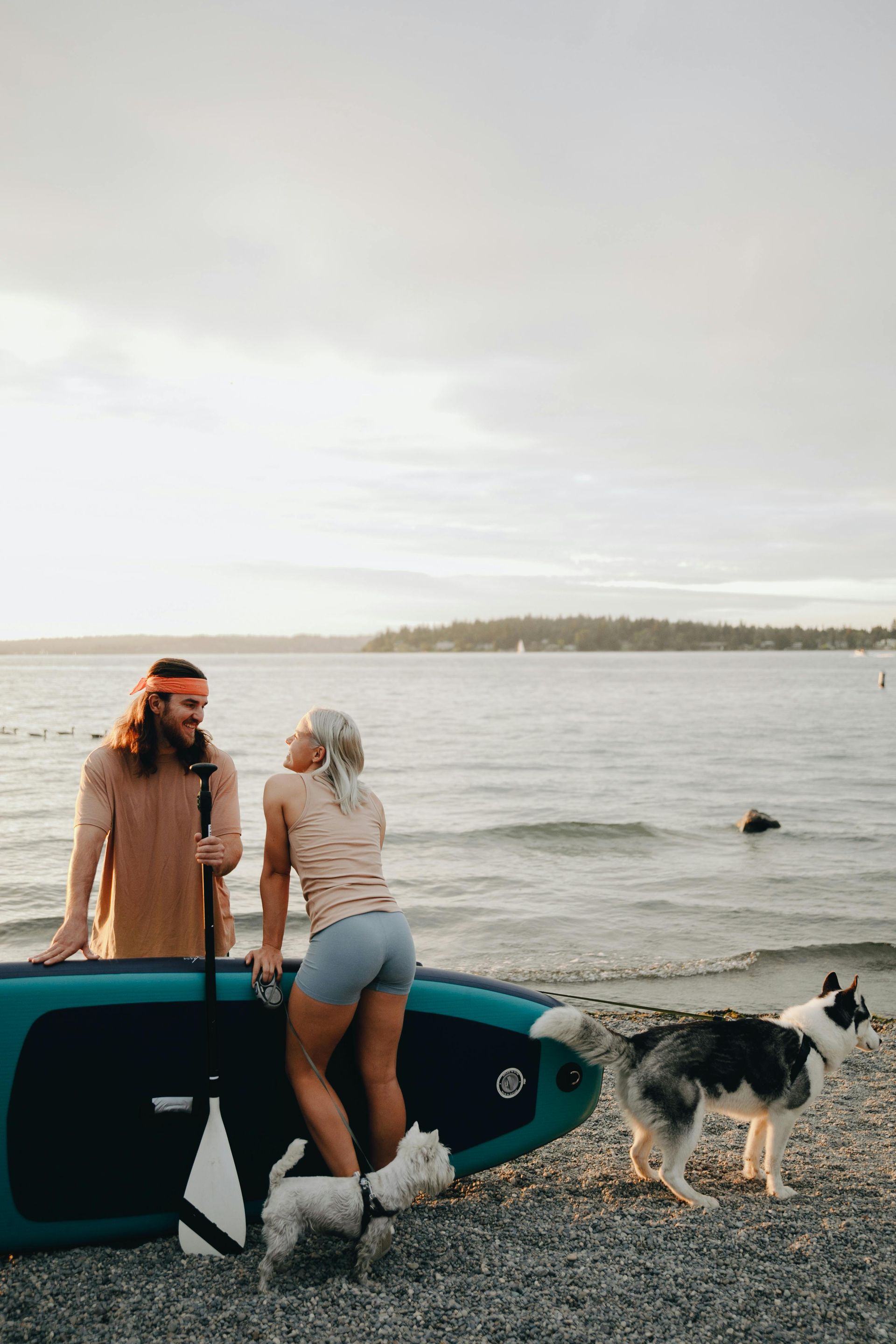 A man and a woman are standing on a beach with a paddle board and a dog.