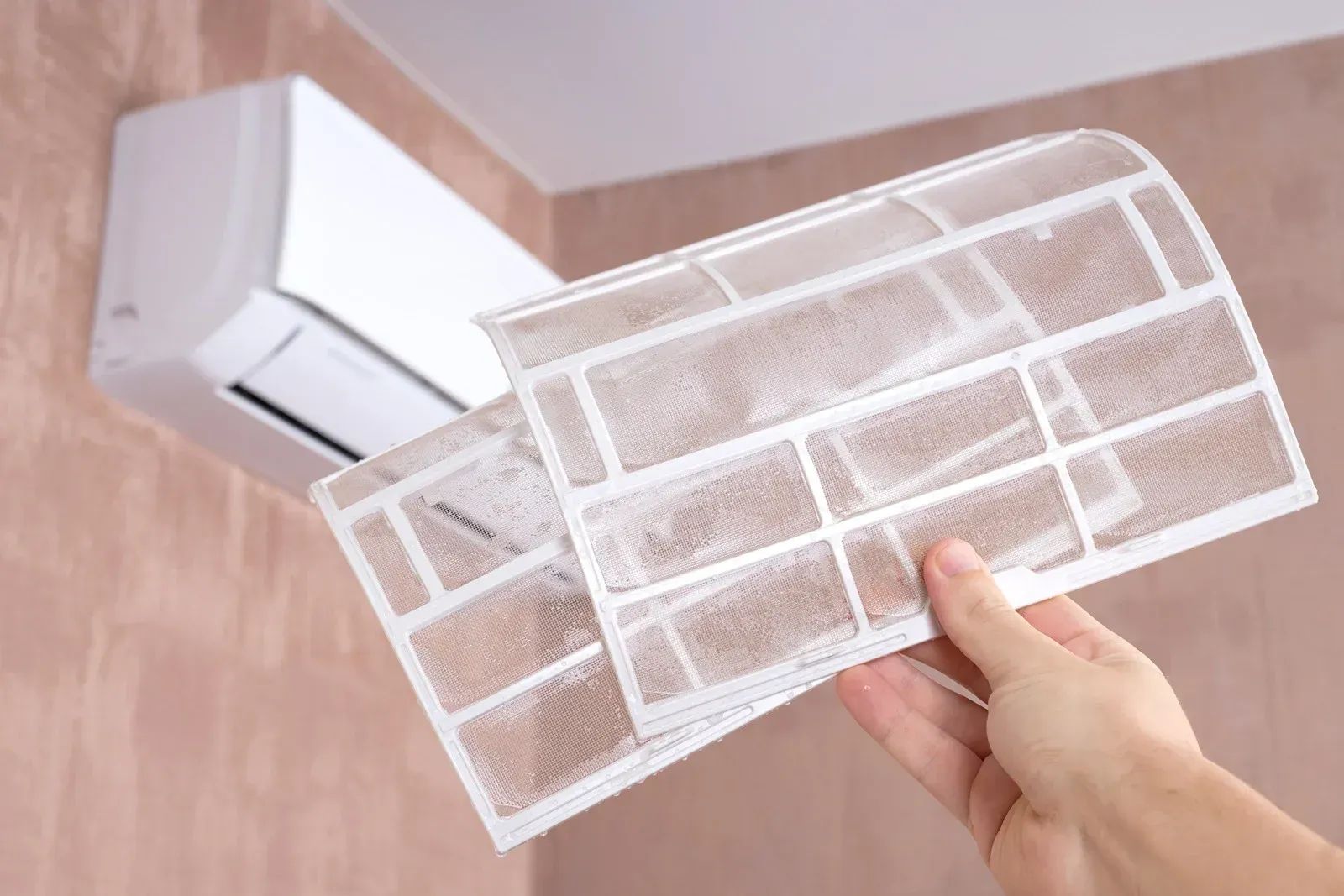 Person holding two dusty air conditioner filters in front of a wall-mounted AC unit.