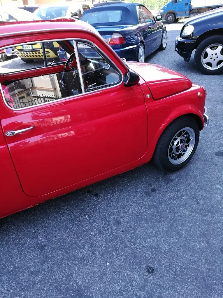 Auto rossa d'epoca parcheggiata sull'asfalto, altri veicoli visibili sullo sfondo.