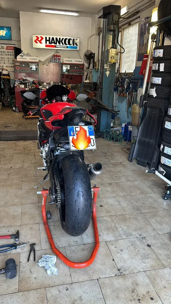 Motocicletta rossa su un supporto arancione all'interno di un'officina disordinata, con l'insegna della Hankook sullo sfondo.