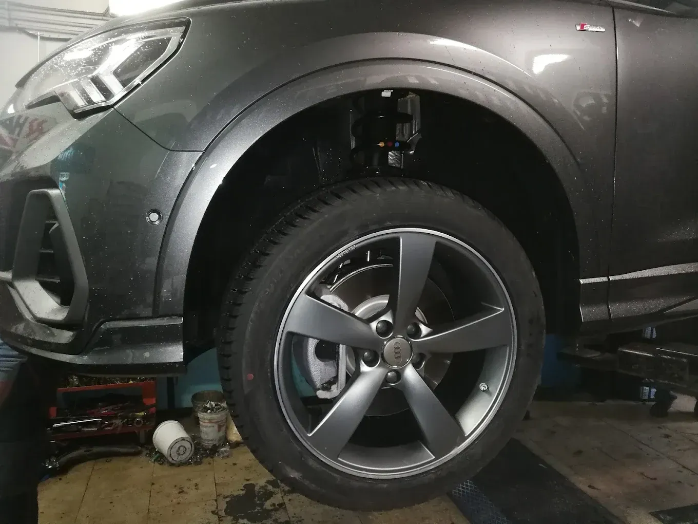 Ruota grigia di un'auto rimossa, che mostra i componenti delle sospensioni e dei freni in un'officina.