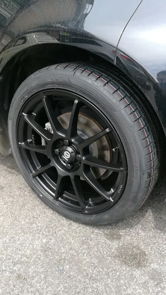 Ruota di un'auto nera con cerchio e pneumatico neri su una superficie grigia.