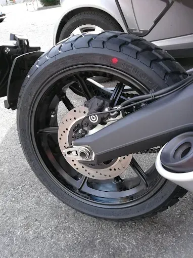 Ruota nera di motocicletta con pneumatico e disco freno; auto grigia sullo sfondo.