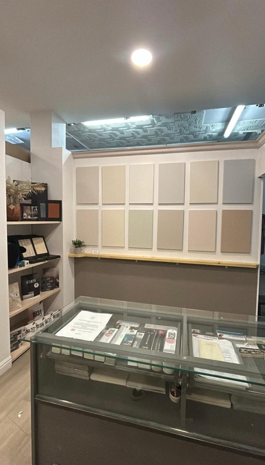 Interno di un negozio caratterizzato da un bancone espositivo in vetro e una parete con dodici campioni quadrati di colore di vernice disposti su due file.