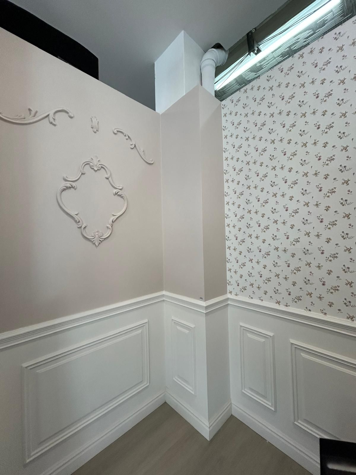Un angolo di stanza con pareti rosa chiaro decorate con modanature e una sezione di carta da parati floreale sopra una boiserie bianca.