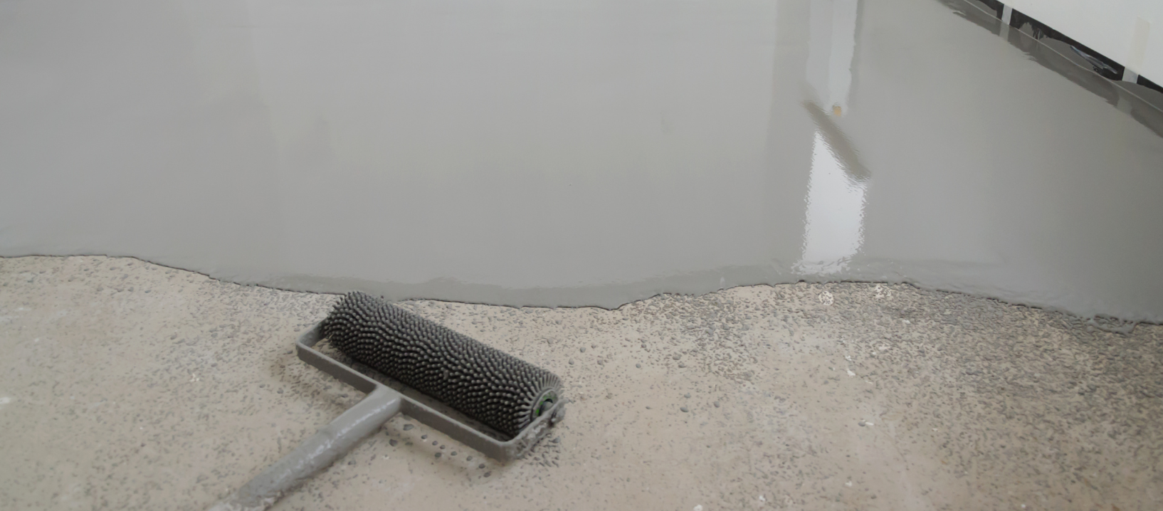 A floor being coated with grey epoxy. A roller lies near the fresh application.