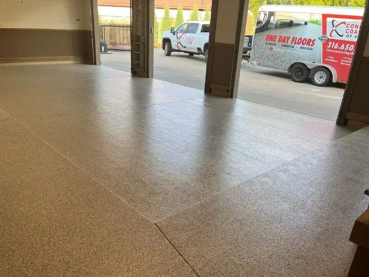 A large room with a newly coated speckled floor, open doorway with a work truck parked outside.