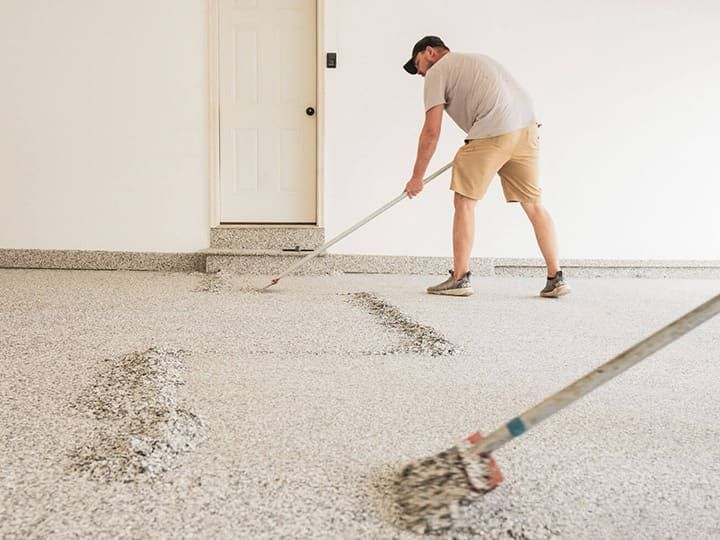 Man using a rake to spread epoxy flakes on a garage floor. White walls, door in the background.