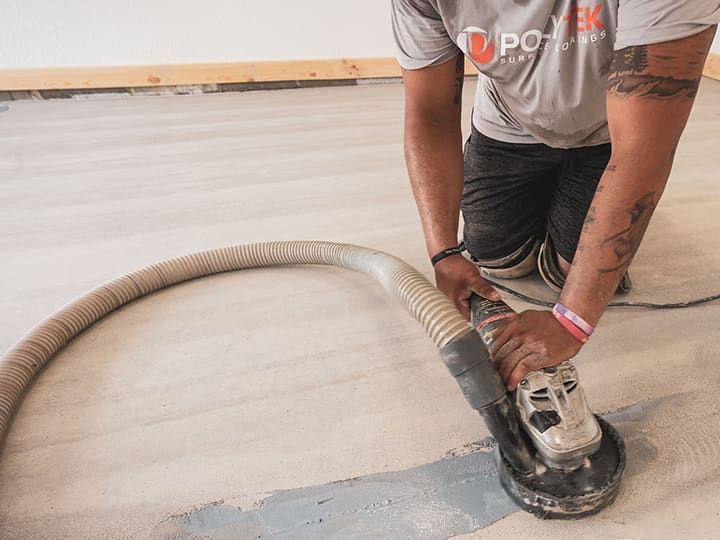 Person grinding a concrete floor with a power tool, wearing a grey shirt and black shorts.