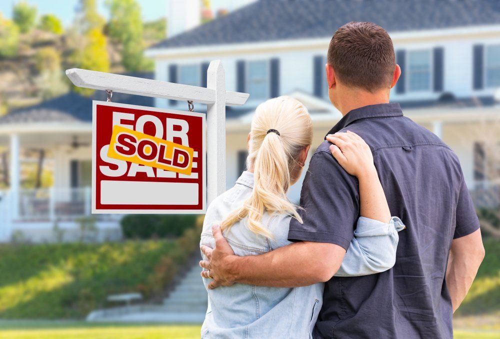 Young Adult Couple Facing Front of Sold Real Estate Sign and House.
