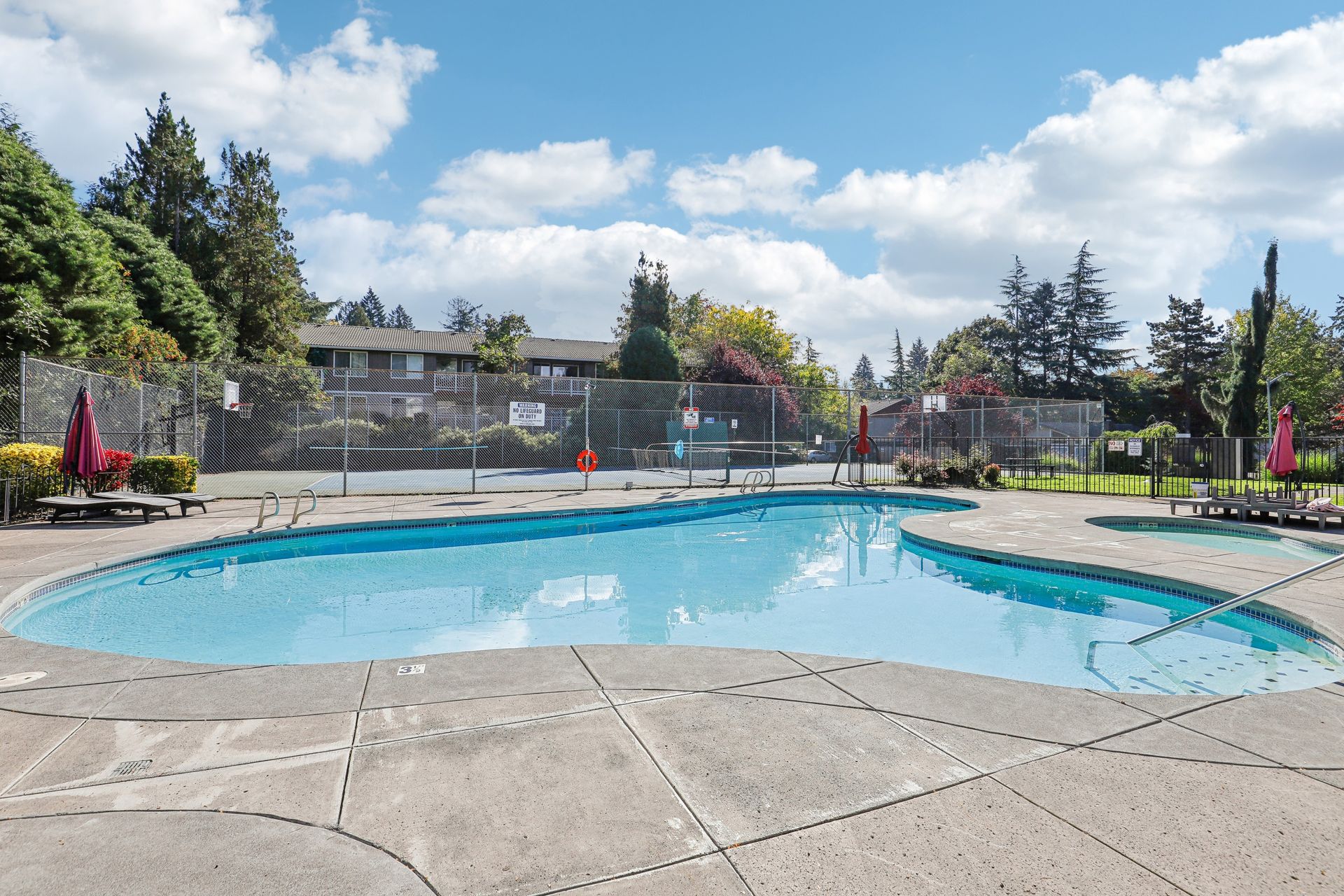 Outdoor community pool with a curved shape, lounge chairs, and fencing under a blue sky.