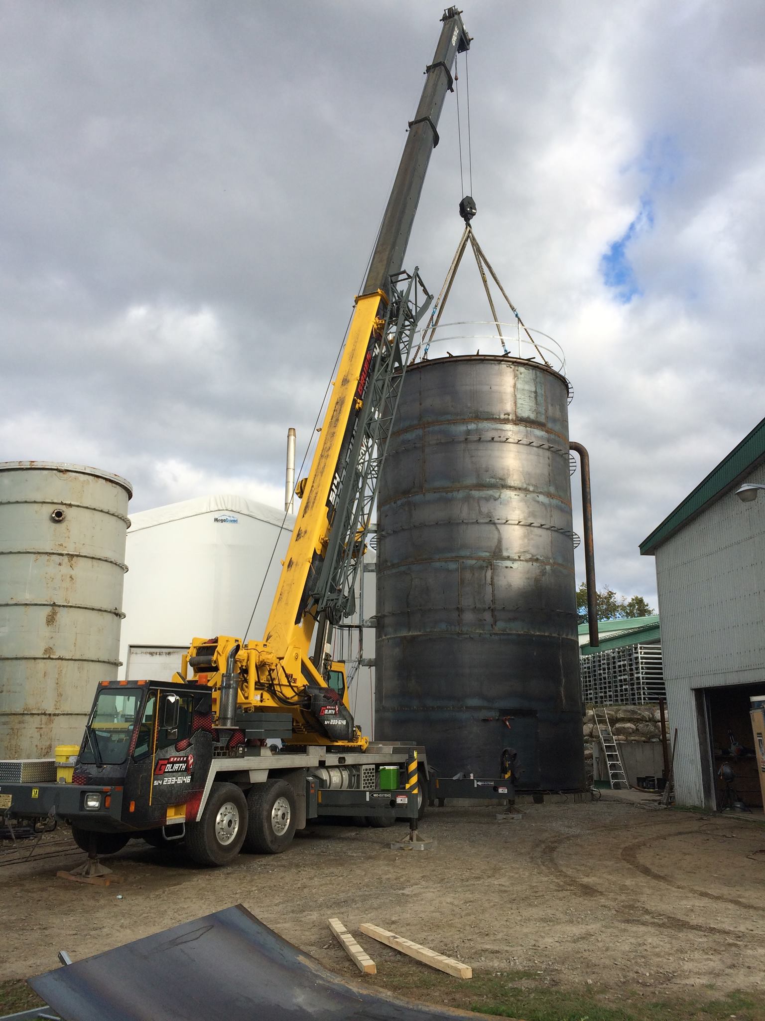 Une grue soulève un grand réservoir cylindrique sombre. Plusieurs autres réservoirs et bâtiments sont visibles à l'arrière-plan.