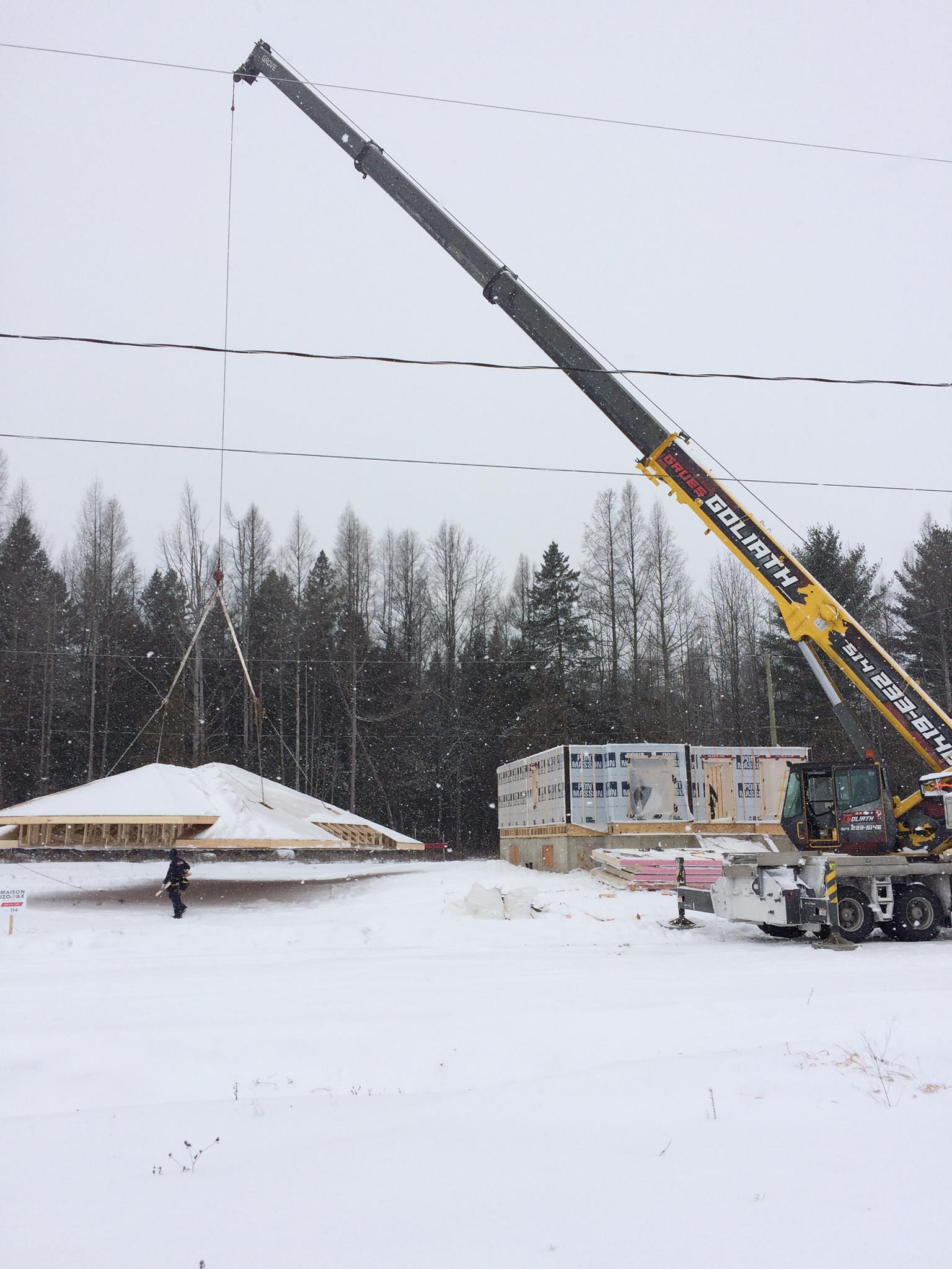 Une grue soulève une ferme de toit au-dessus d'un chantier de construction enneigé, avec des arbres en arrière-plan.