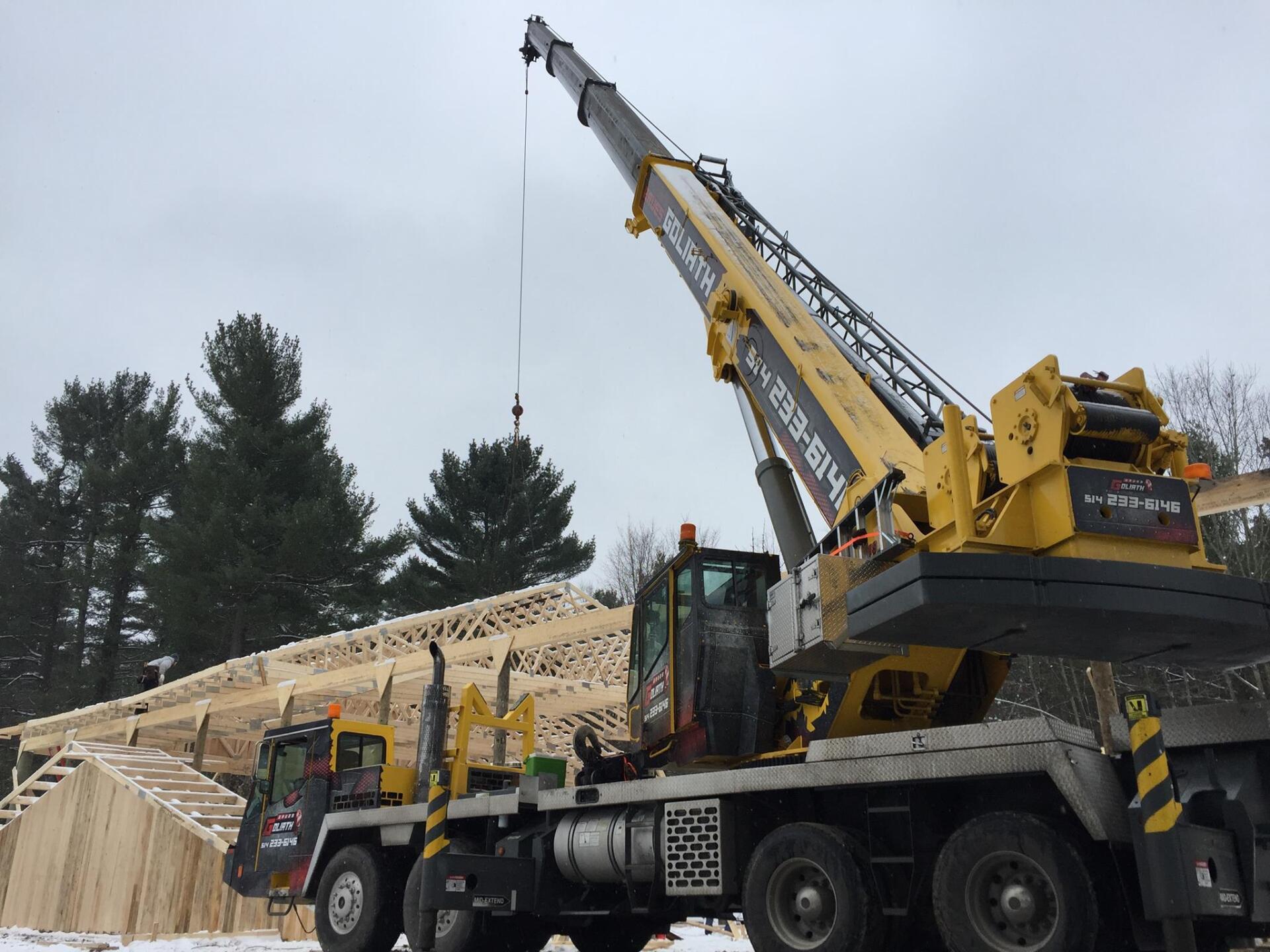 Grue jaune soulevant des fermes de toit en bois sur un chantier de construction ; décor extérieur enneigé.