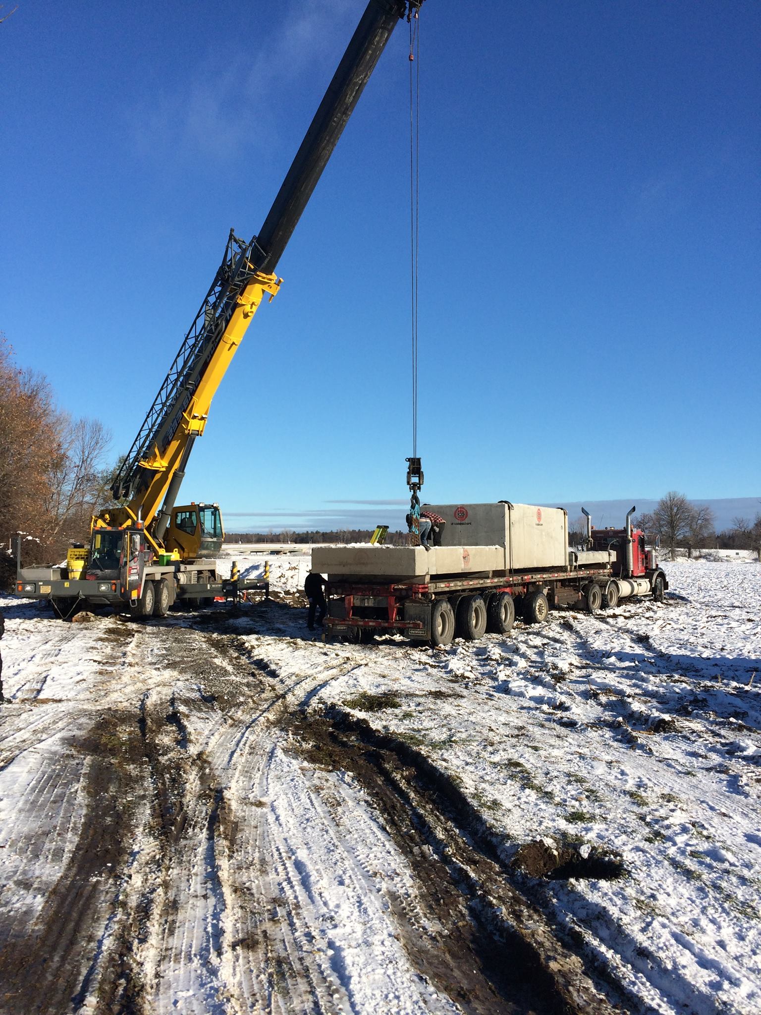 Grue soulevant une structure rectangulaire d'un camion à plateau dans un champ enneigé par une journée ensoleillée.