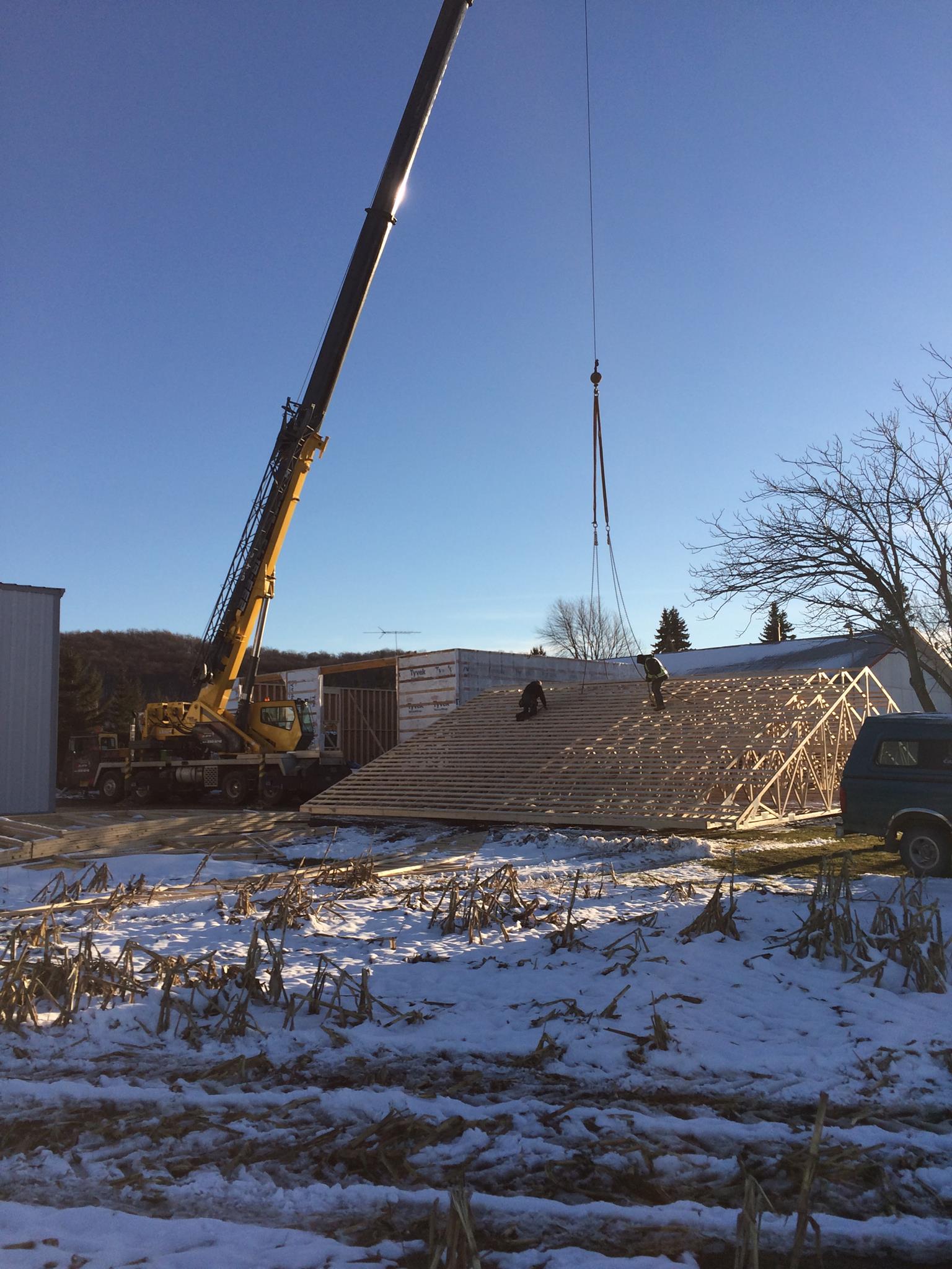 Une grue soulève une section de toit sur un bâtiment en construction par une journée enneigée.
