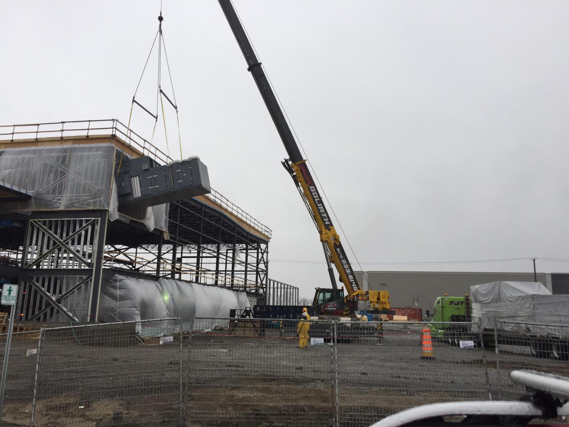 Une grue soulève une grande unité de chauffage, de ventilation et de climatisation grise sur un bâtiment en construction. Journée nuageuse.