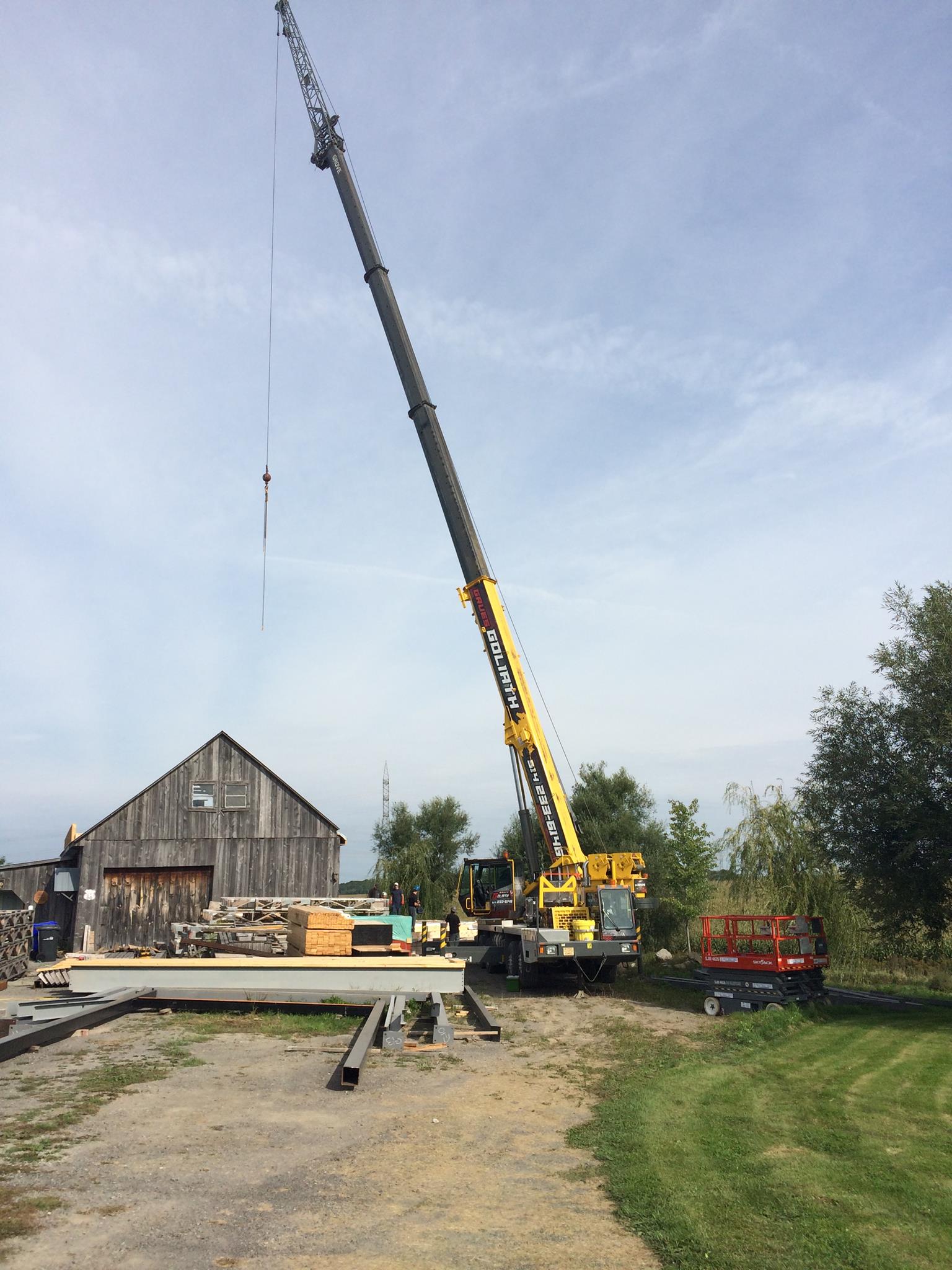 Grue soulevant des poutres près d'une grange délabrée et d'équipements sur un terrain en gravier. Ciel bleu.