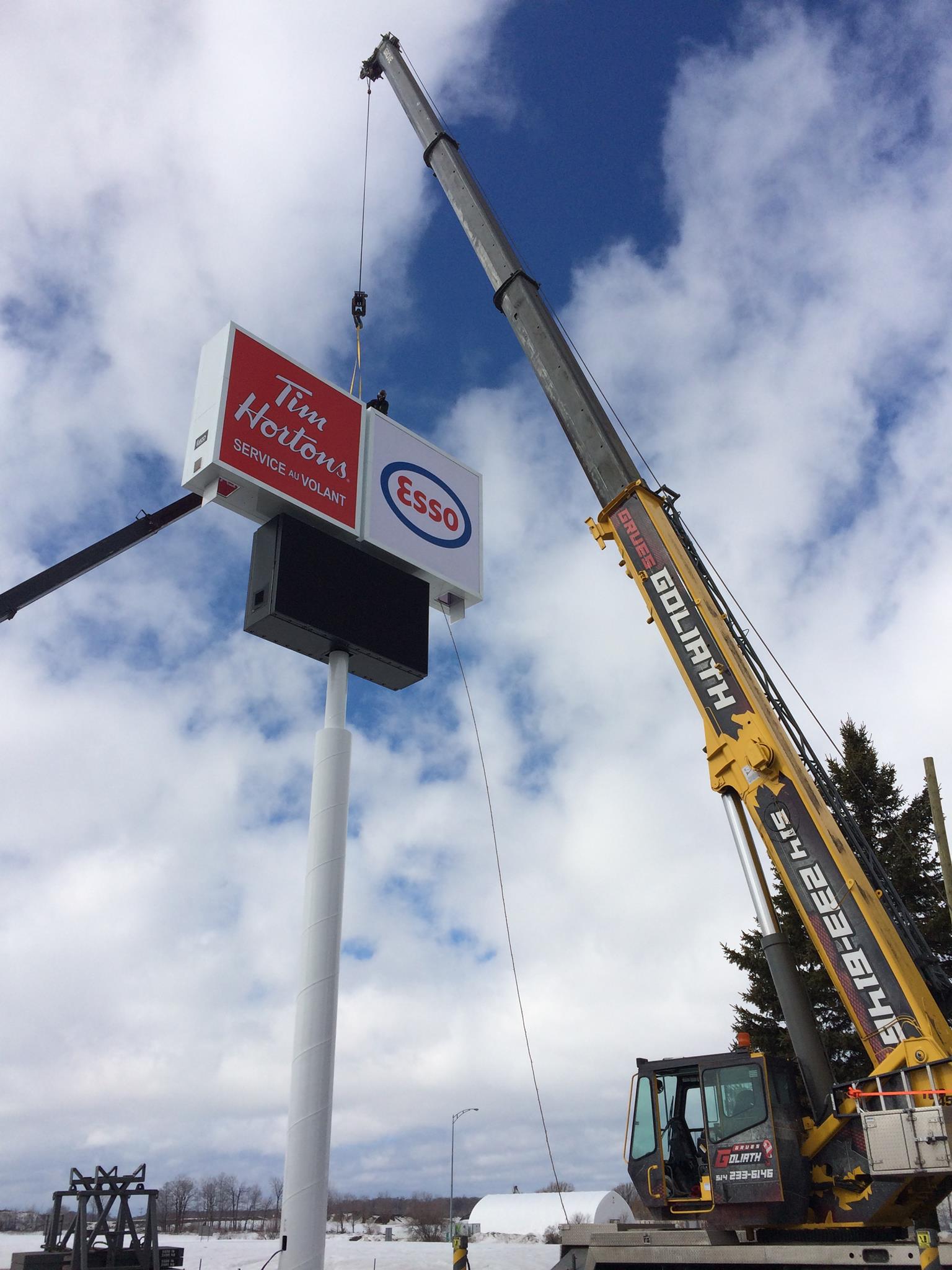 Une grue soulève une enseigne Tim Hortons et Esso sur un grand poteau blanc contre un ciel nuageux.