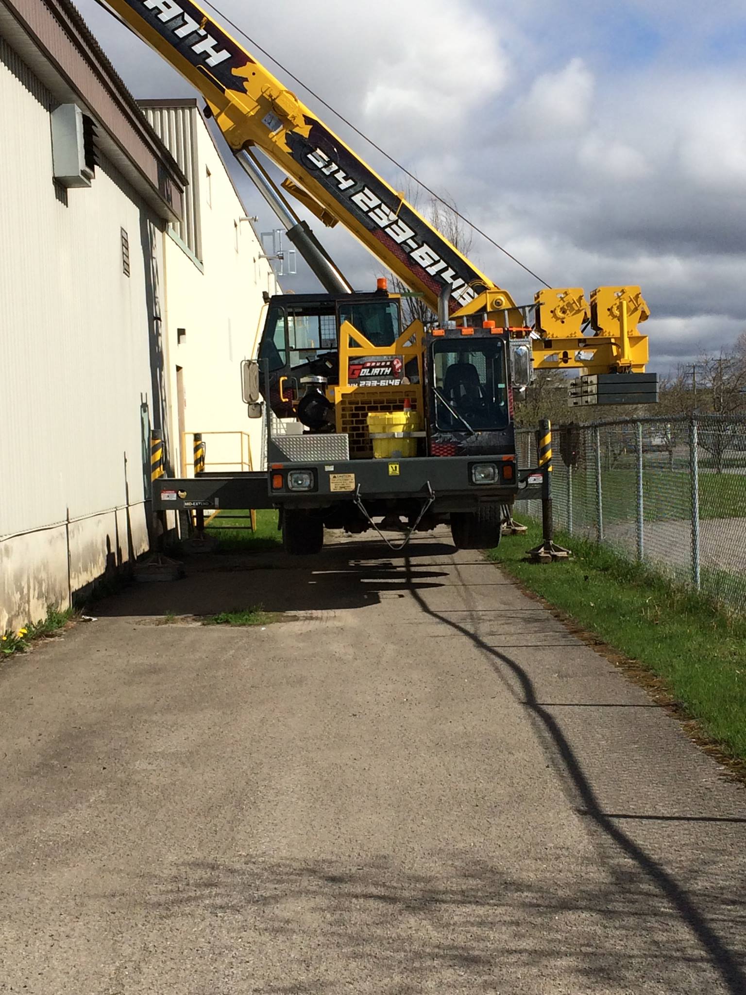 Grue jaune à côté d'un bâtiment sur un chemin en béton ; ciel nuageux en arrière-plan.