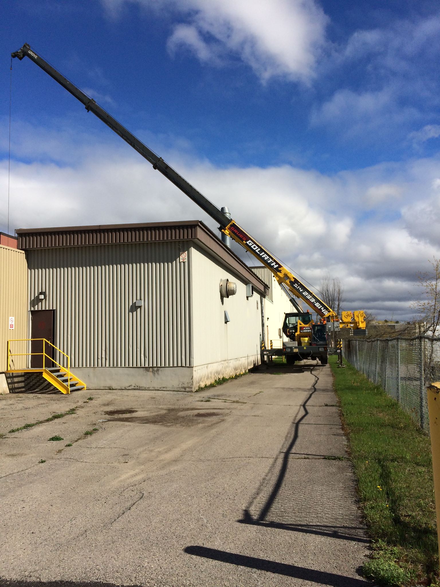 Une grue jaune surplombe un bâtiment blanc au toit marron. Une personne ou un objet est soulevé.