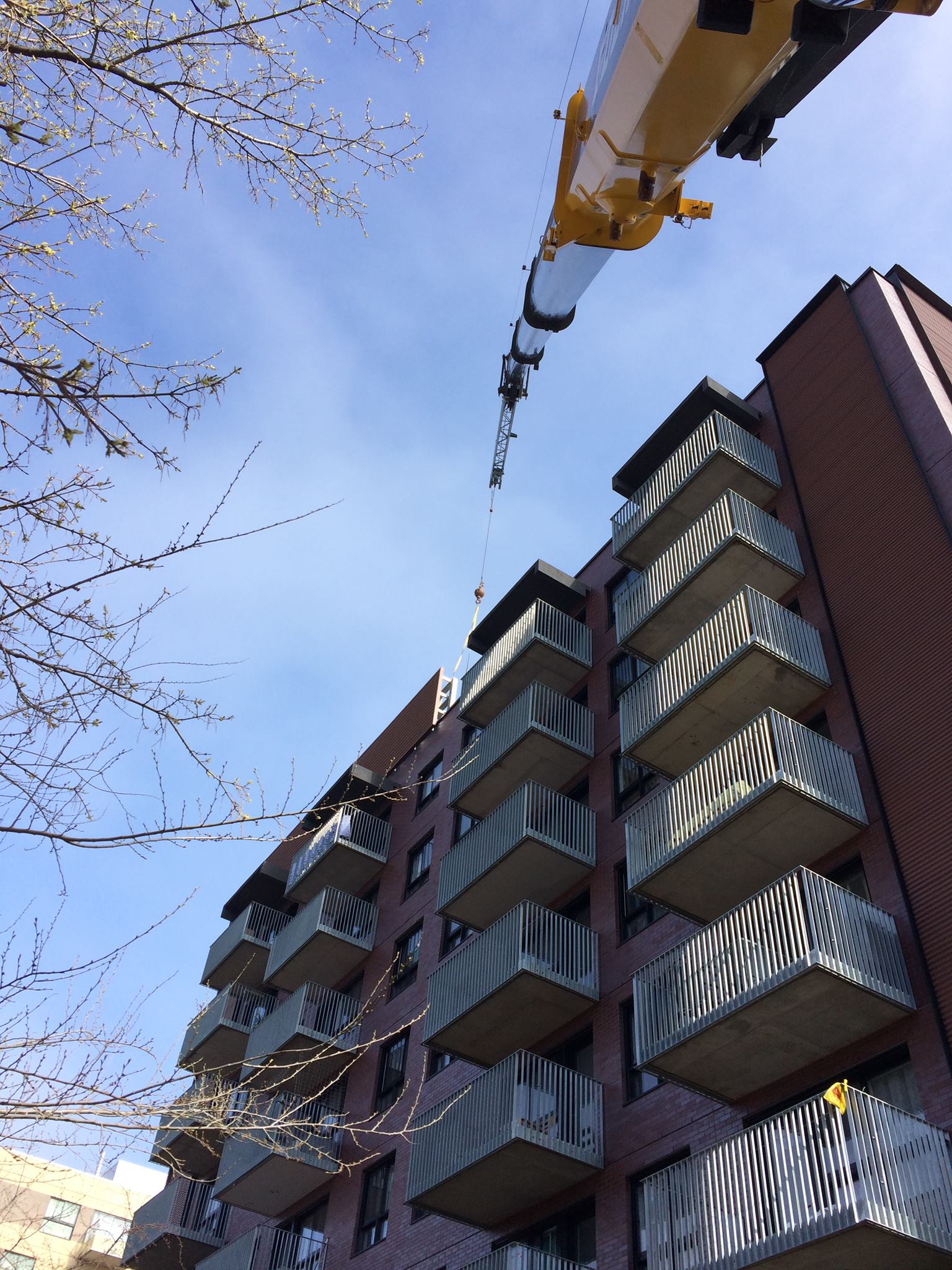 Une grue soulève un objet sur le toit d'un immeuble en briques. Des balcons bordent le bâtiment ; le ciel est bleu.