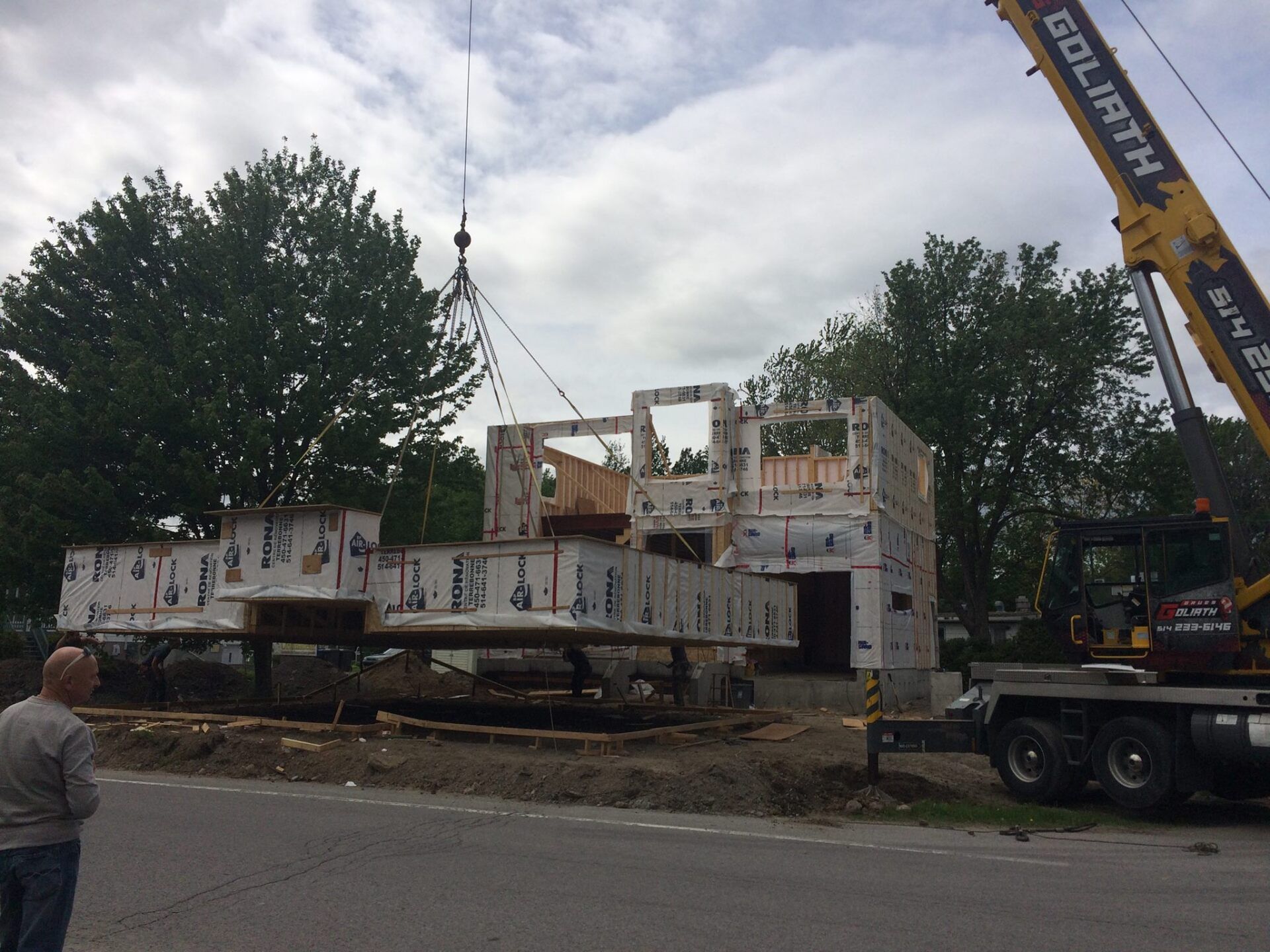 Une grue soulève un module de maison sur un chantier de construction à côté d'une route ; un homme regarde.