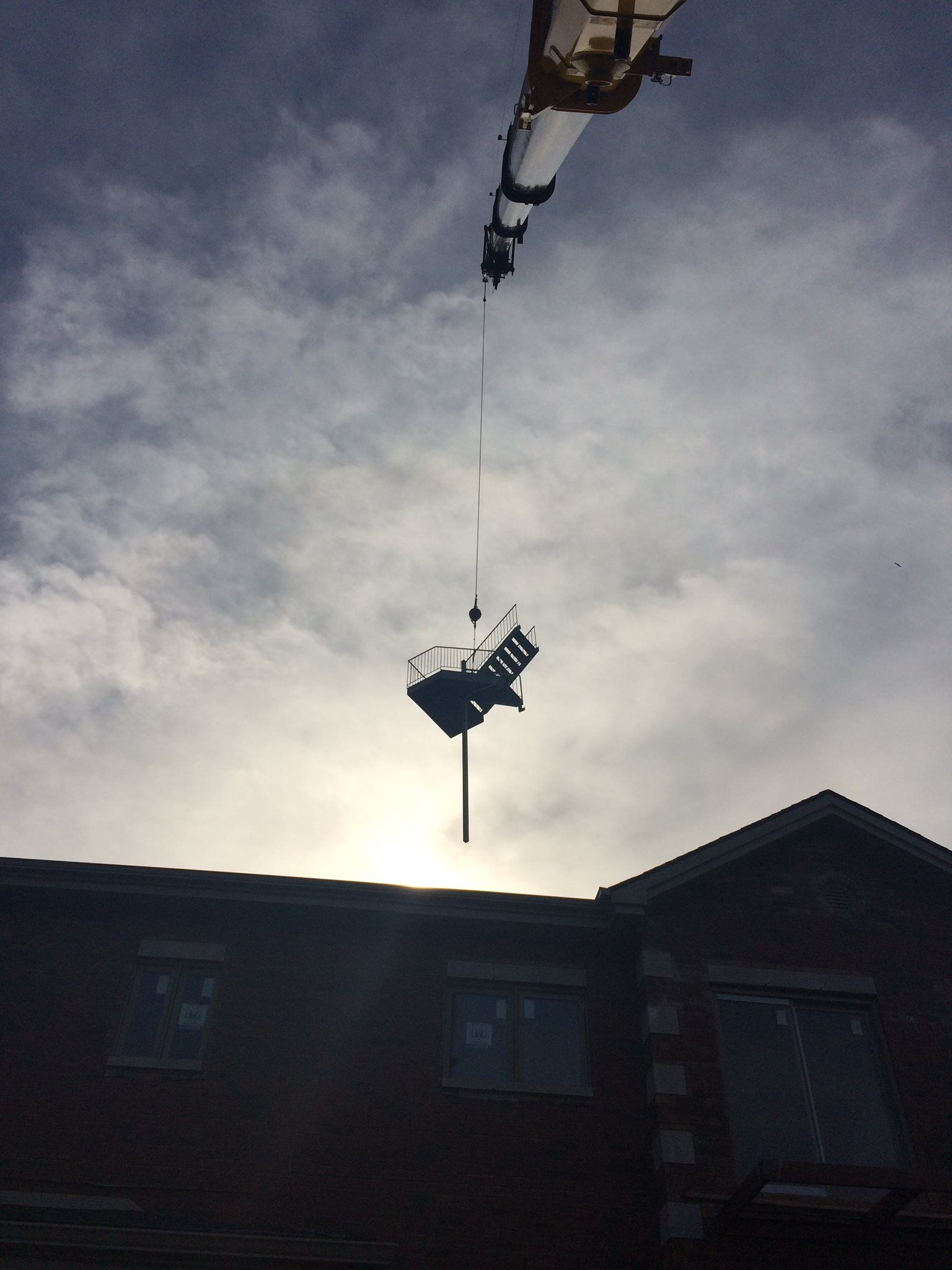 Grue soulevant une structure métallique au-dessus d'un bâtiment en briques contre un ciel nuageux.