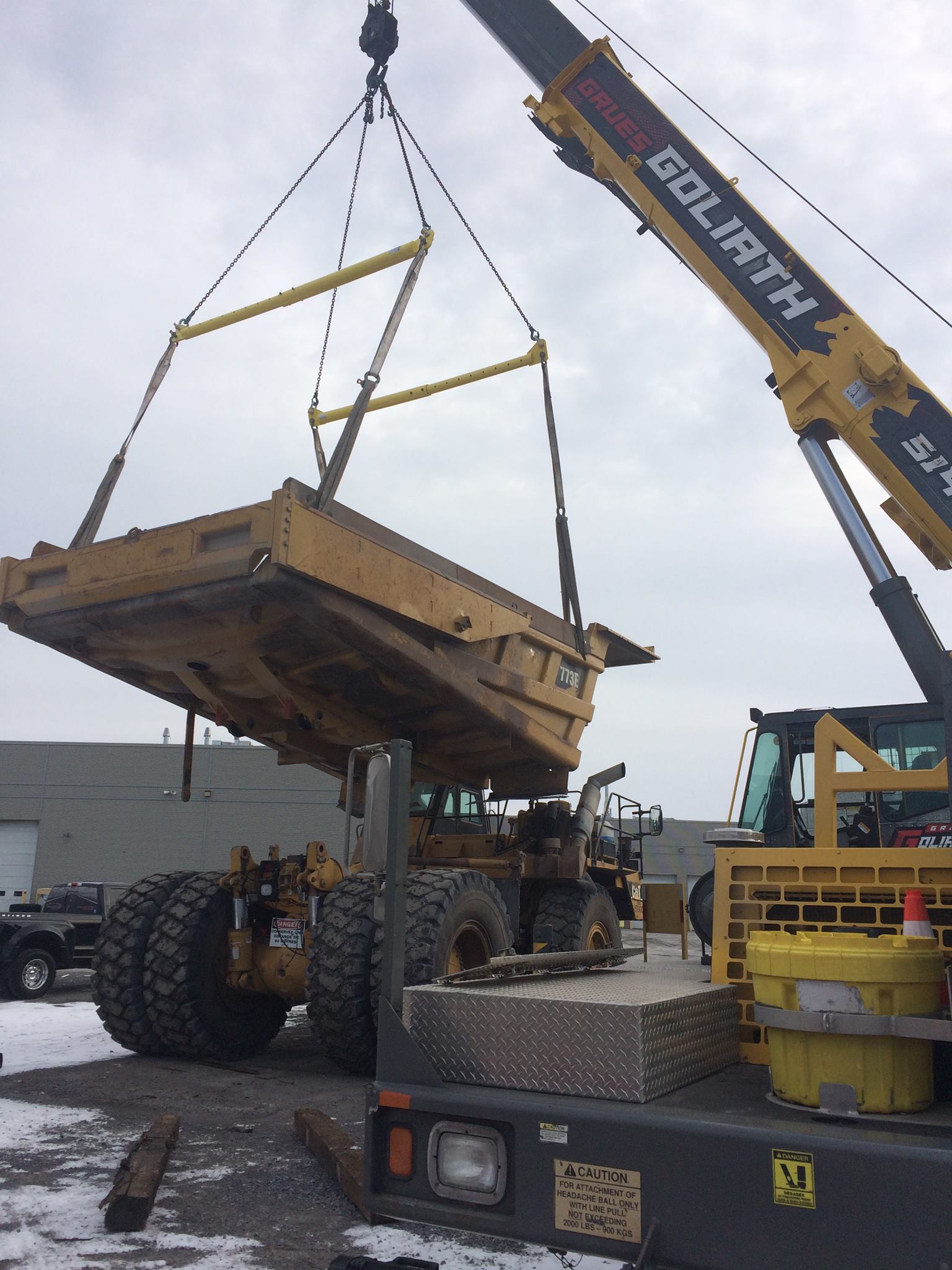 Une grande benne de camion à benne basculante jaune soulevée par une grue jaune « Goliath » sur une benne de camion, à l'extérieur.