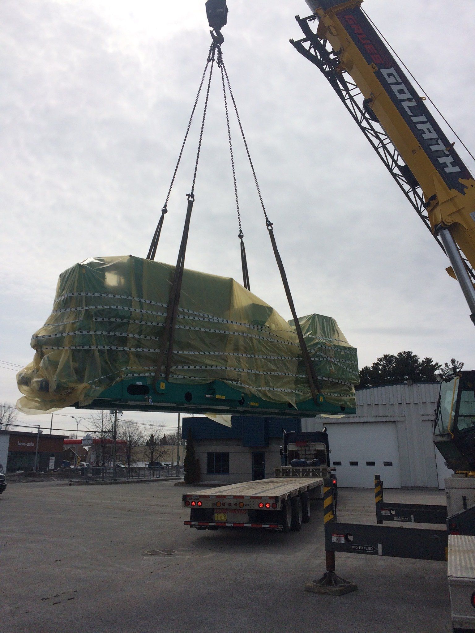 Une grue soulève une grande machine verte au-dessus d'un camion à plateau, à l'extérieur par une journée nuageuse.