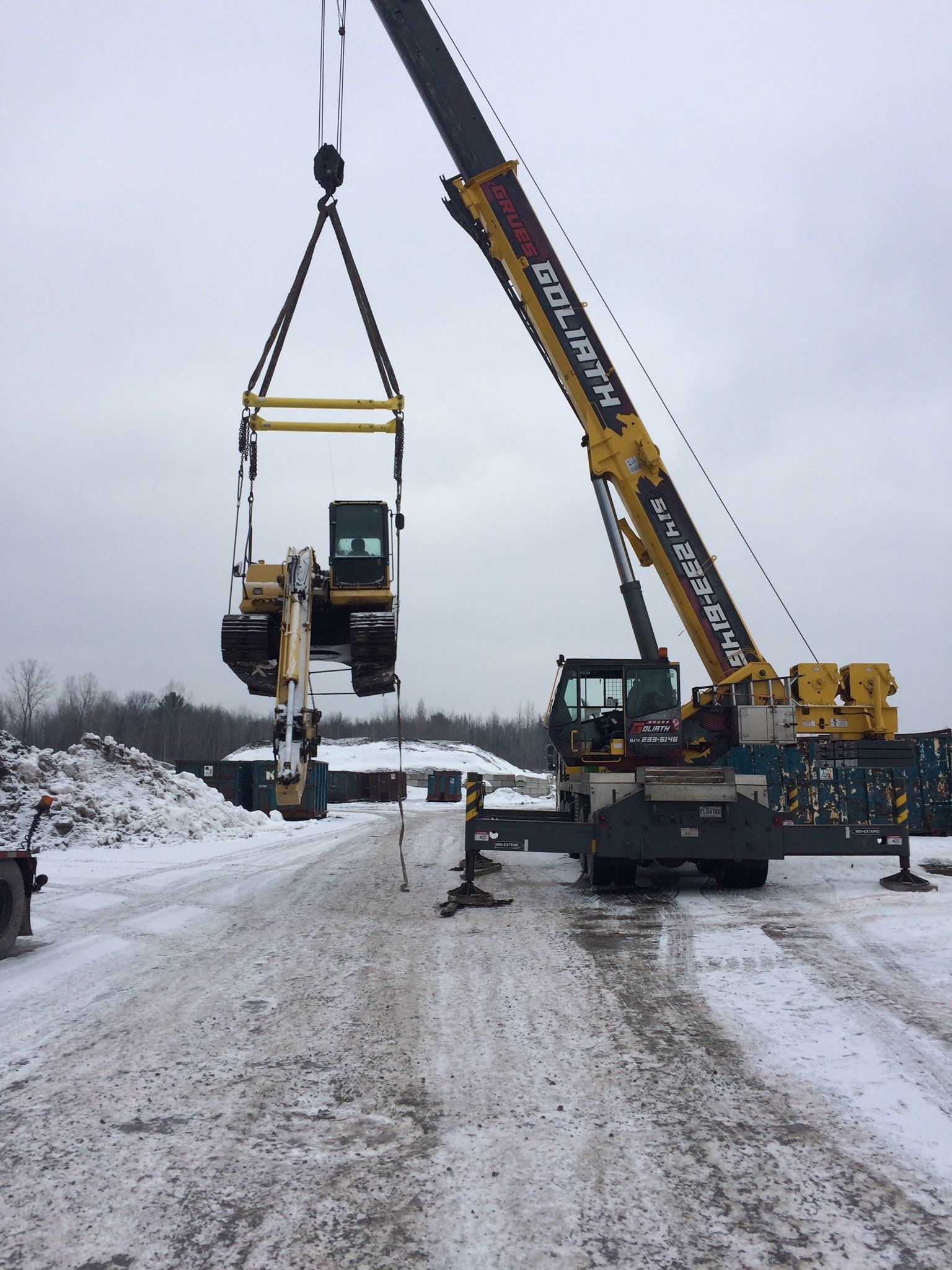 Une grue soulevant une excavatrice d'une route enneigée par temps nuageux.