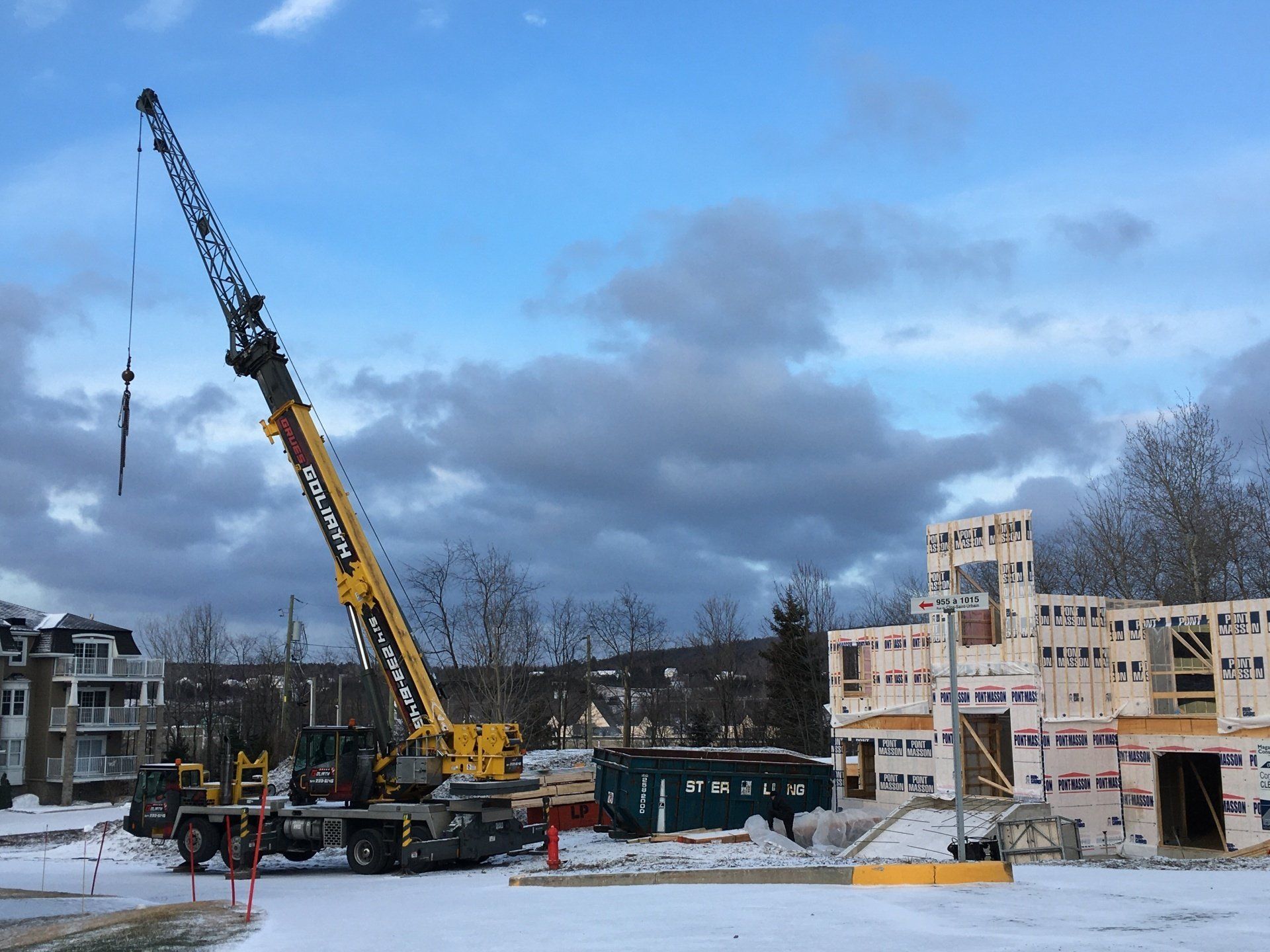 Chantier de construction avec grue soulevant des matériaux ; charpente en bois d'un bâtiment en construction. Neige au sol.