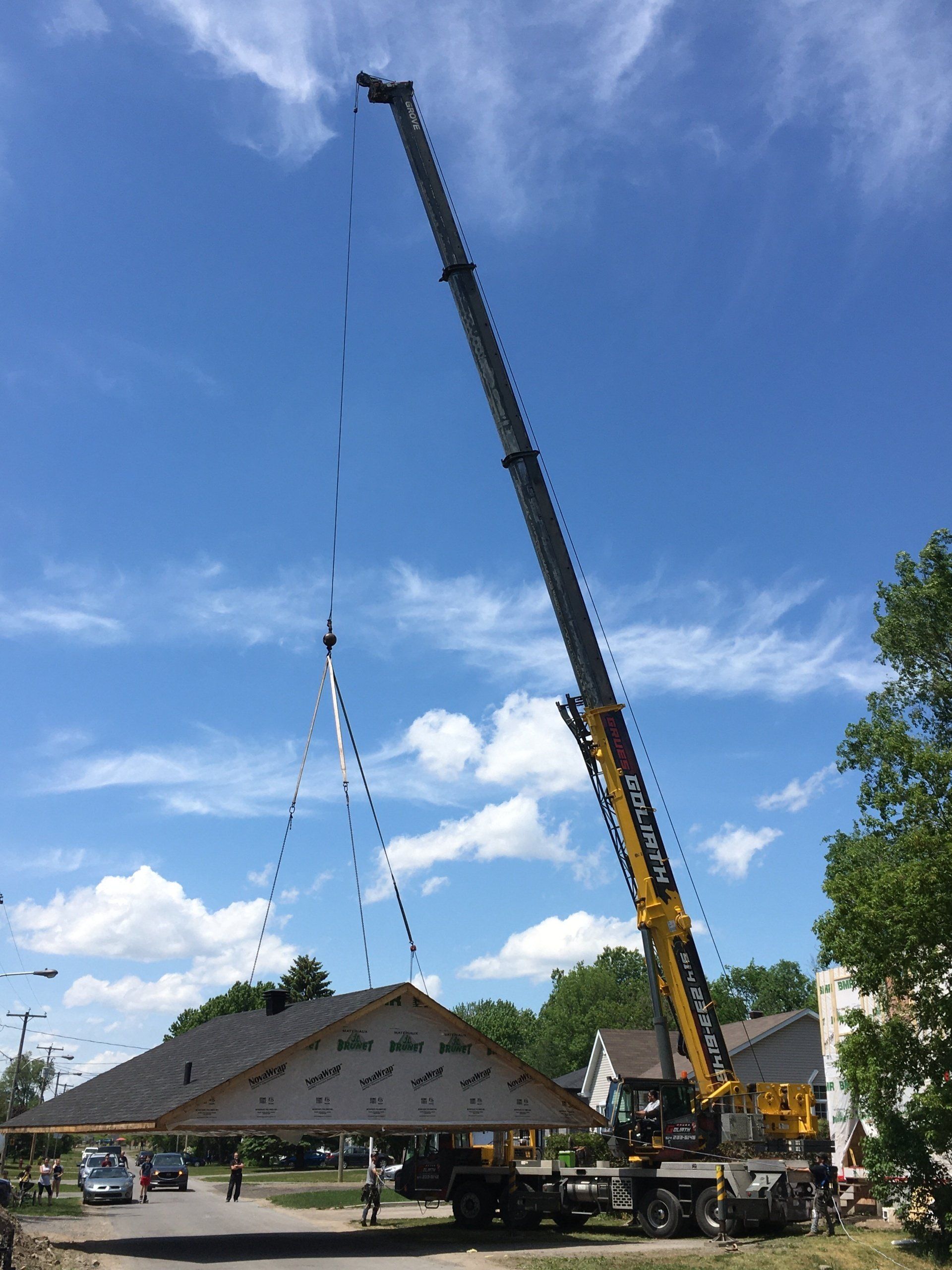 Une grue soulève une grande section de toit au-dessus d'une rue résidentielle par une journée ensoleillée.