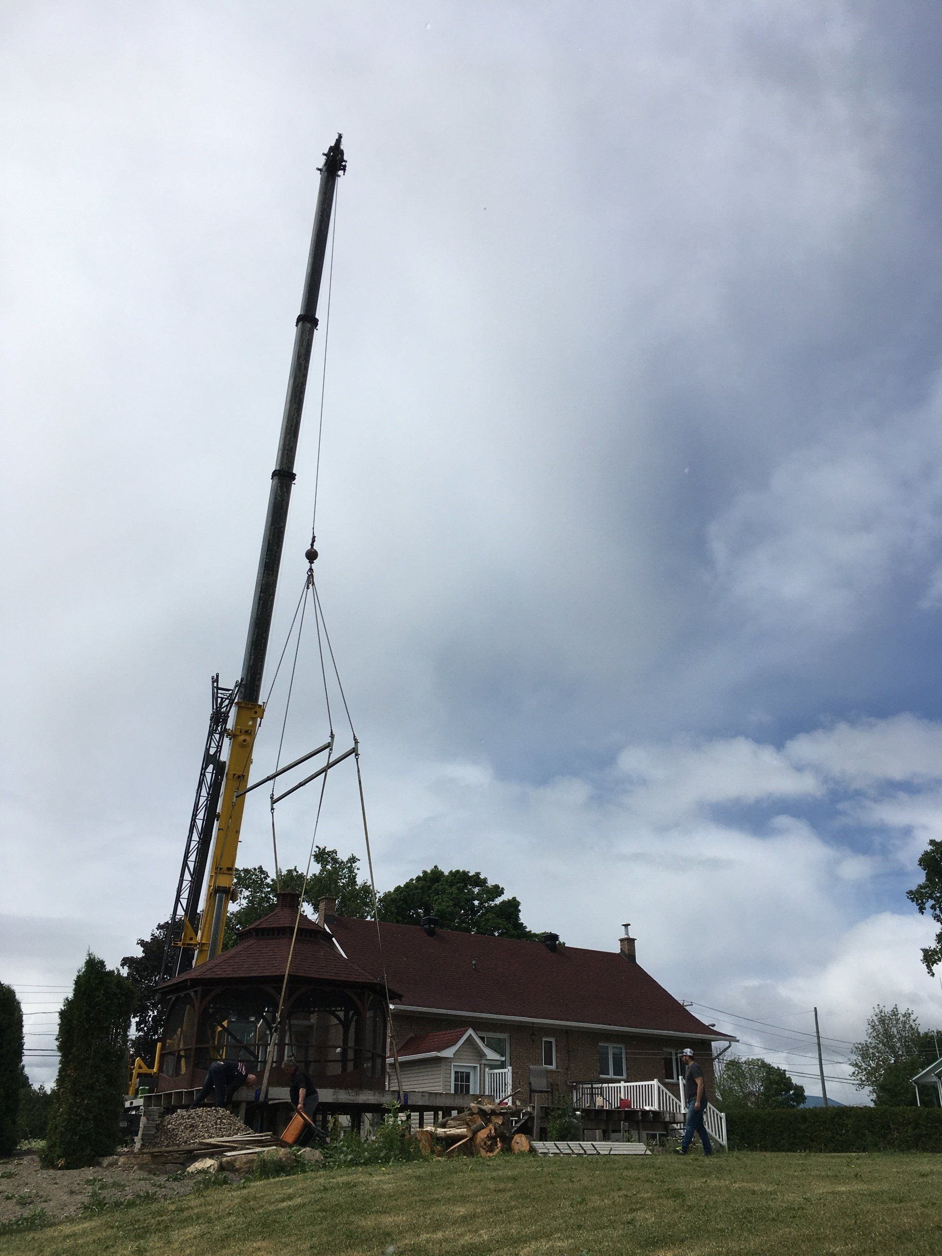 Une grue soulève une structure de belvédère sur le toit d'une maison à deux étages sous un ciel nuageux.
