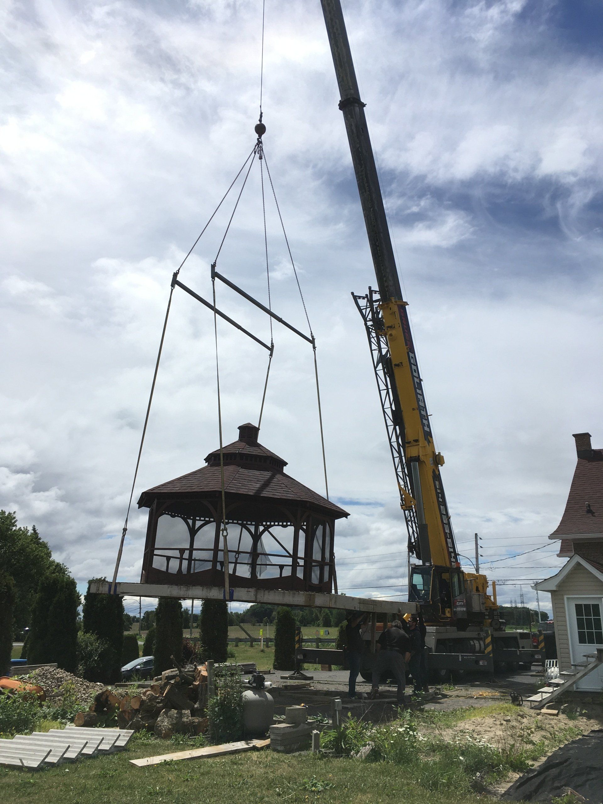 Une grue soulève un belvédère dans une cour. Ciel lumineux et petite maison en arrière-plan.